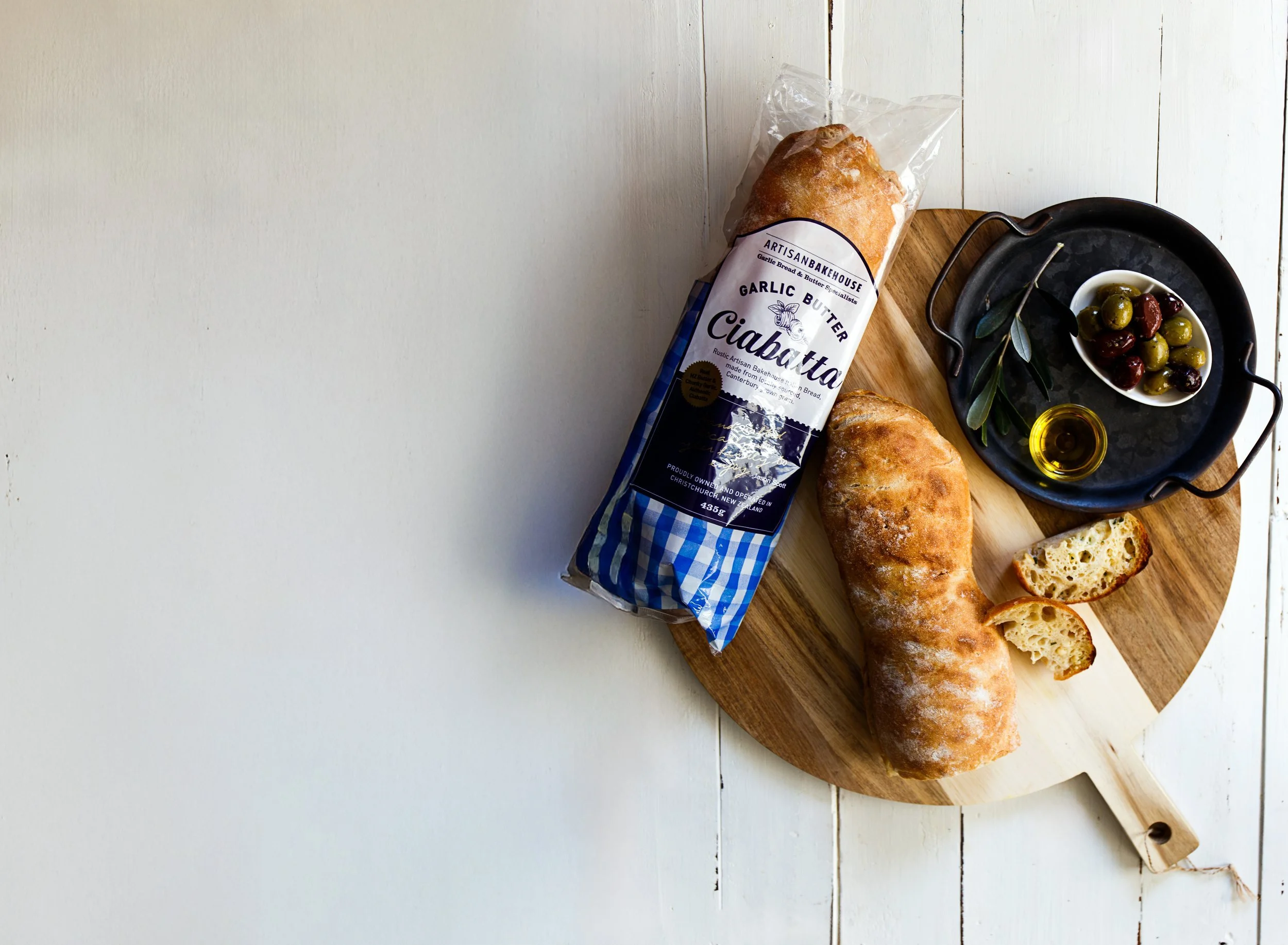 A loaf of garlic bread in plastic wrapping on a wooden cutting board beside a small plate of mixed olives, some leaves, and a small bottle of olive oil on a white wooden surface.