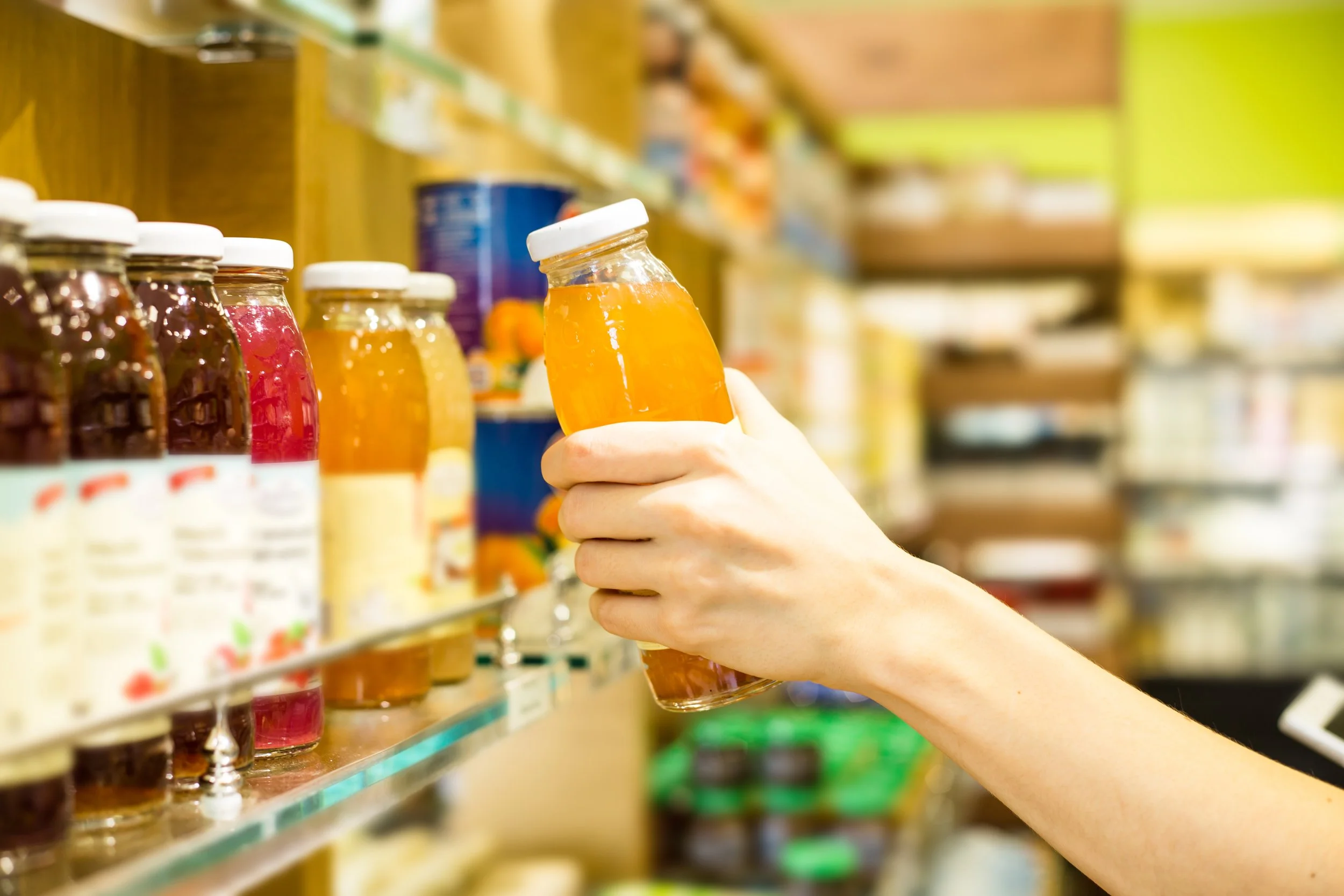 A person holding a small glass jar filled with orange-colored liquid, in a grocery store aisle with various other jars and products on shelves.