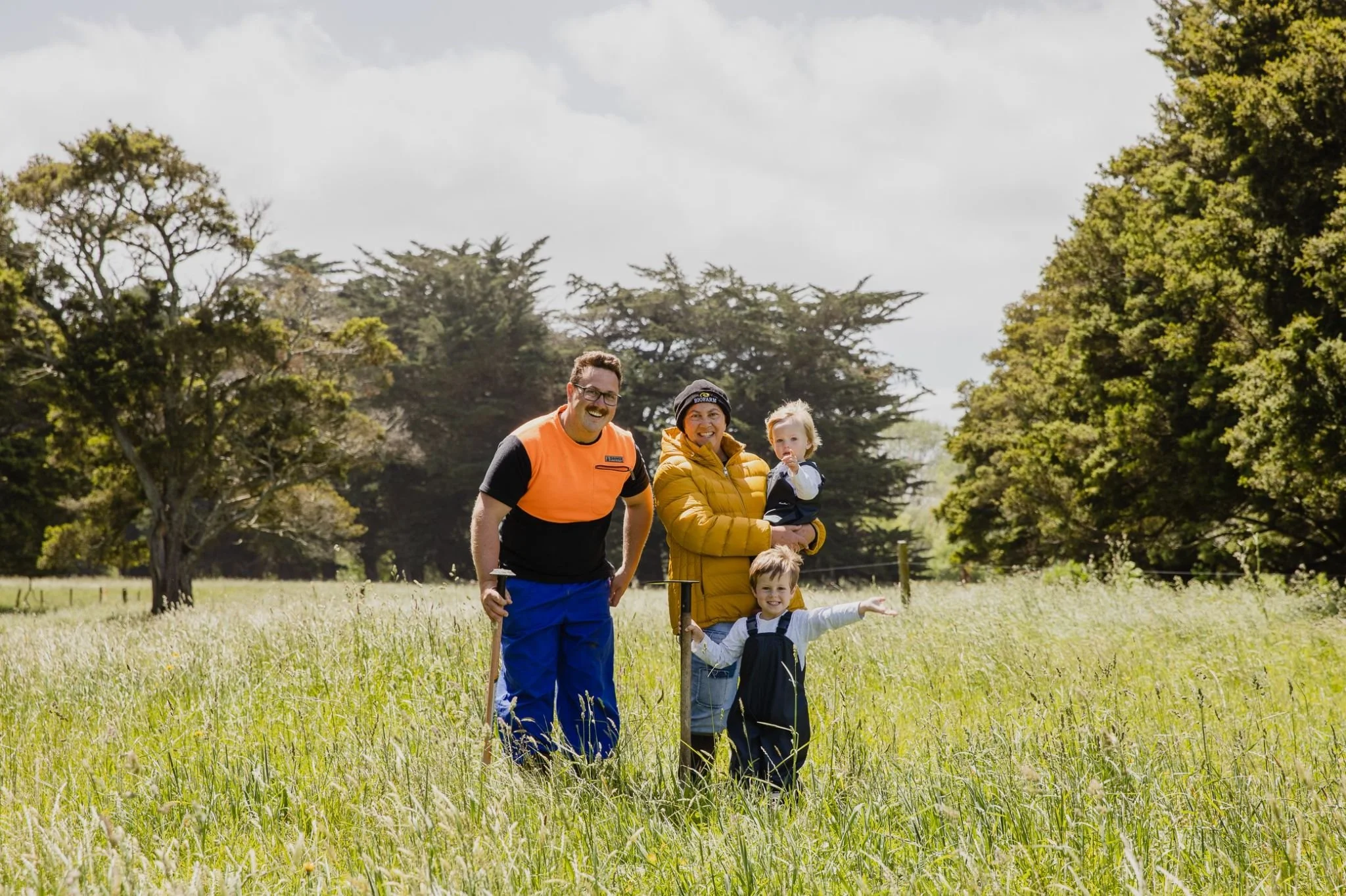 A family of four, including two children and two adults, standing in a grassy field surrounded by tall trees with a cloudy sky overhead, enjoying an outdoor activity together.
