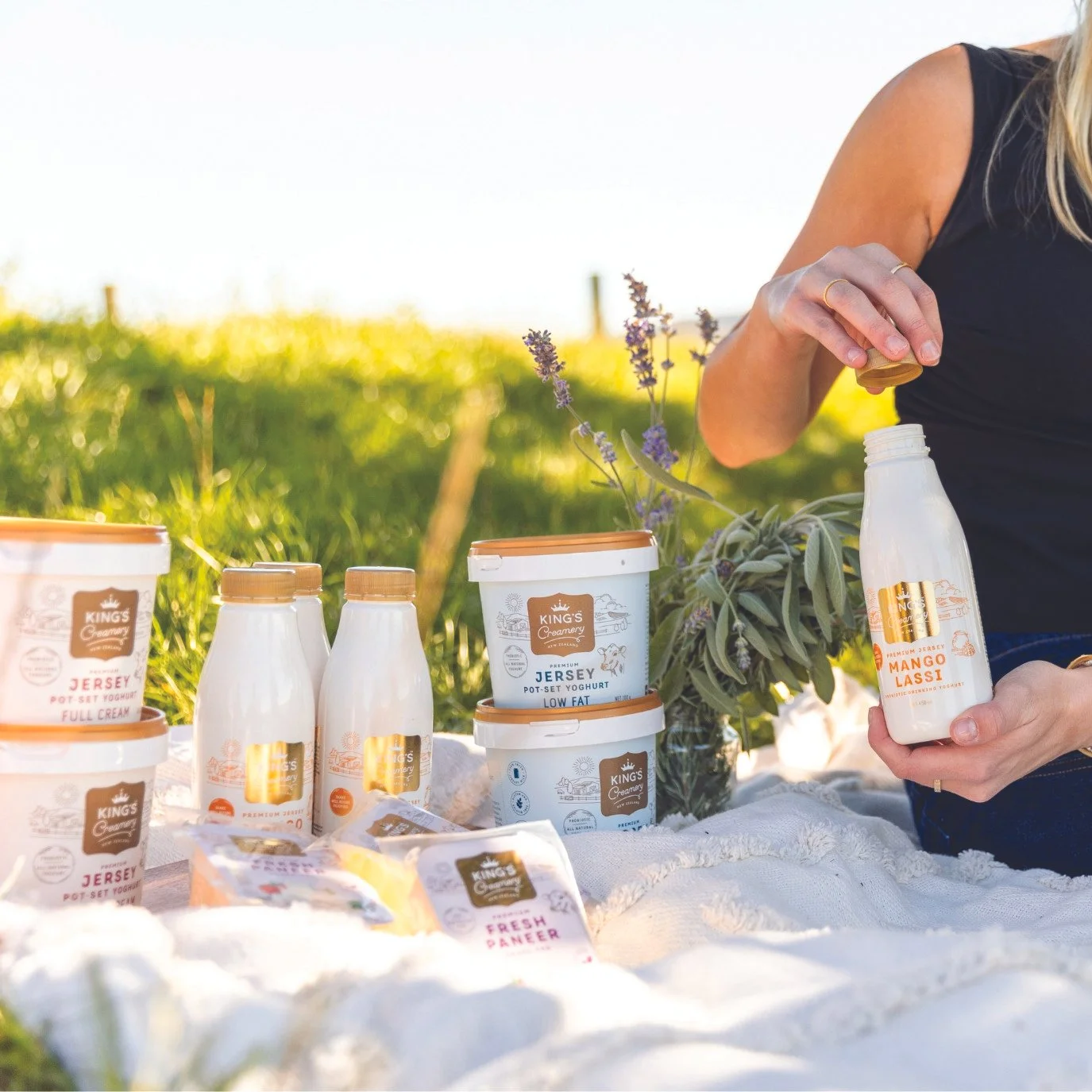 A woman outdoors on a sunny day is pouring yogurt from a bottle, with various yogurt containers, small bottles, and a jar of lavender on a white cloth in front of her.