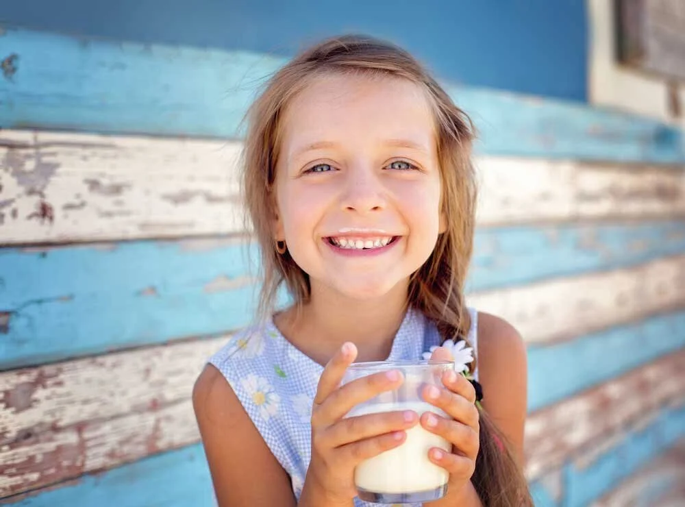 A young girl with light brown hair and blue eyes smiling, holding a glass of milk outside in front of a weathered, painted wooden wall.