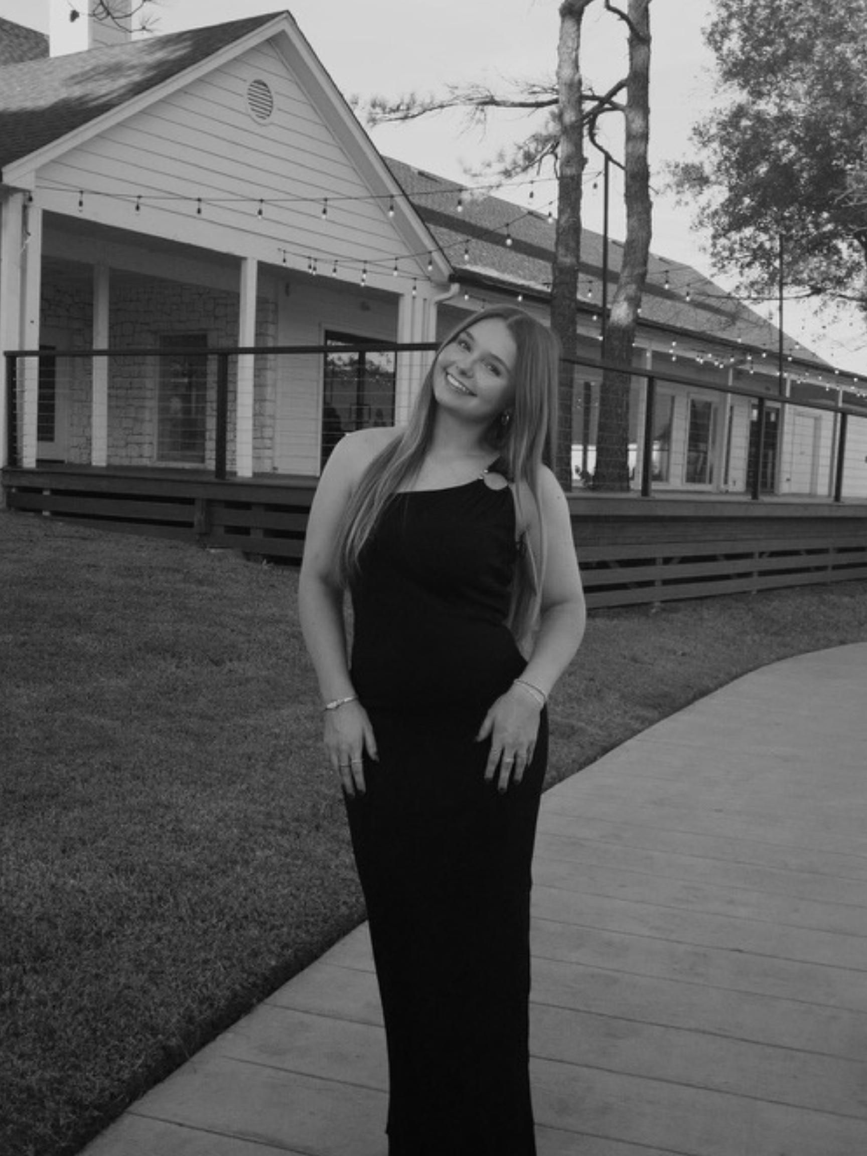 A smiling young woman in a black strapless dress posing outdoors in front of a house with string lights, trees, and a wooden deck.