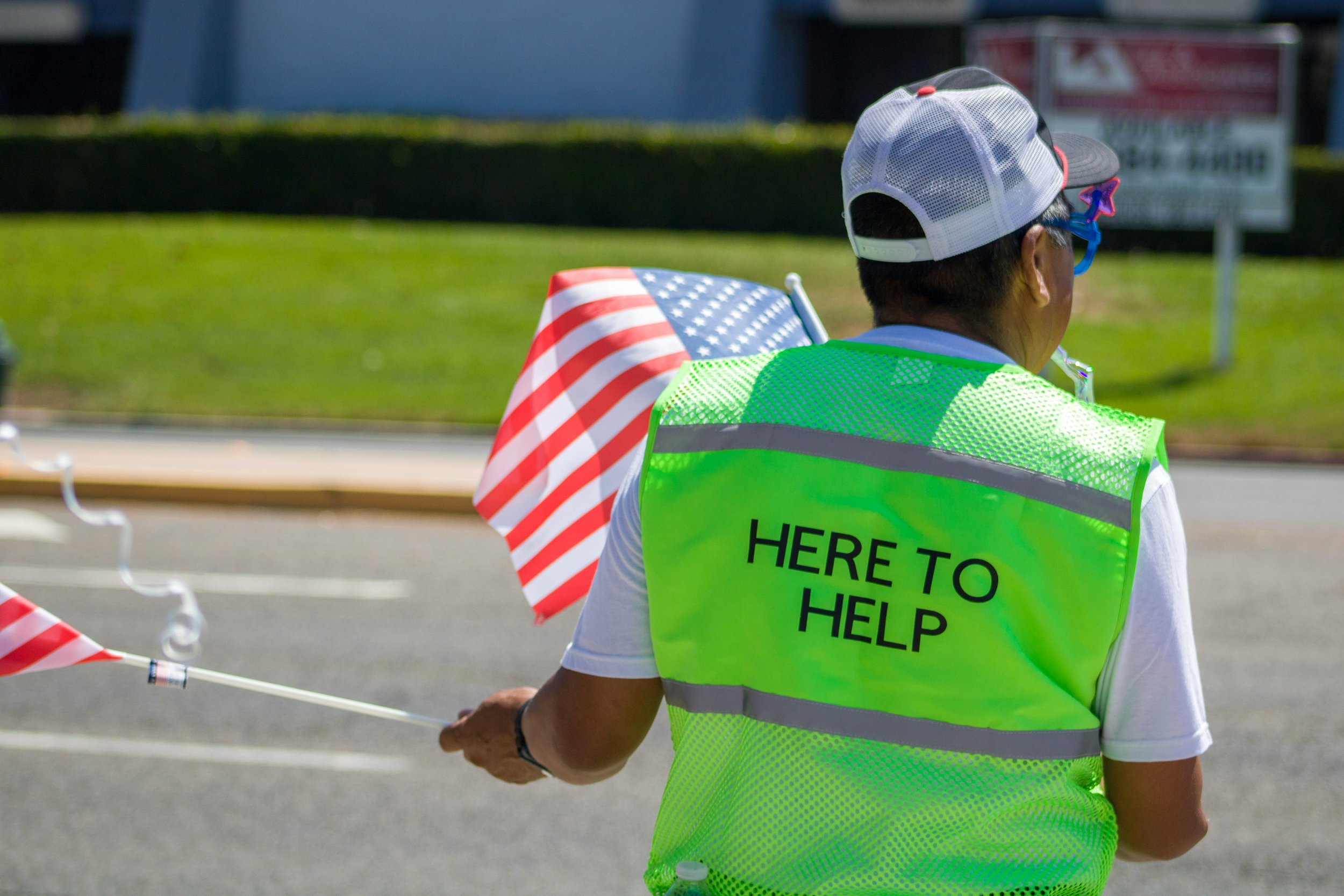 Person wearing a bright yellow vest that says 'Here to Help,' holding an American flag during an outdoor event on a sunny day.