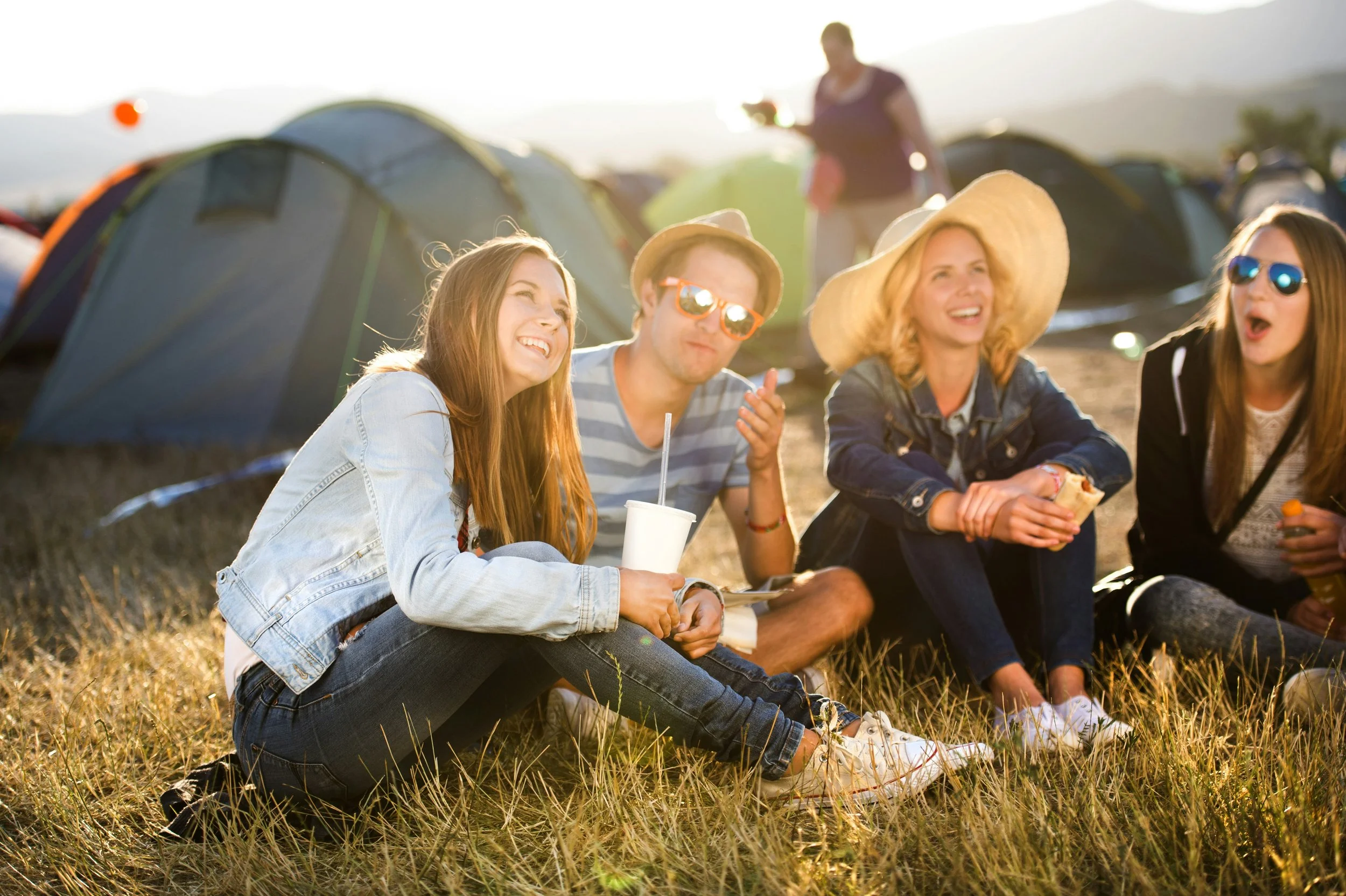 Group of four friends sitting on grass near tents, enjoying outdoor activity in late afternoon sunlight, with one person wearing a wide-brim hat and others sunglasses and casual clothing.