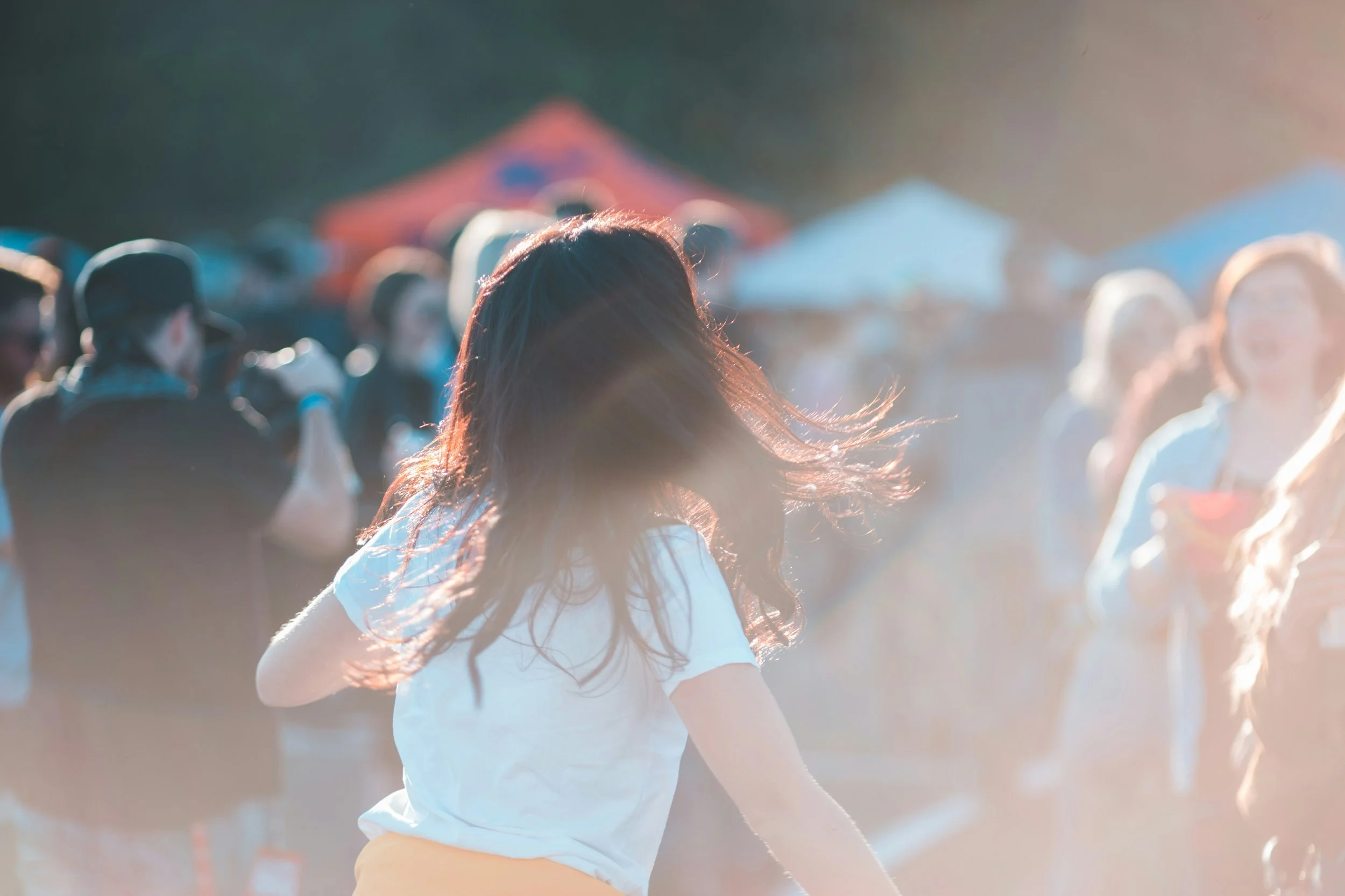 A woman with long hair wearing a white T-shirt running among a crowd at sunset with a blurred background of tents.