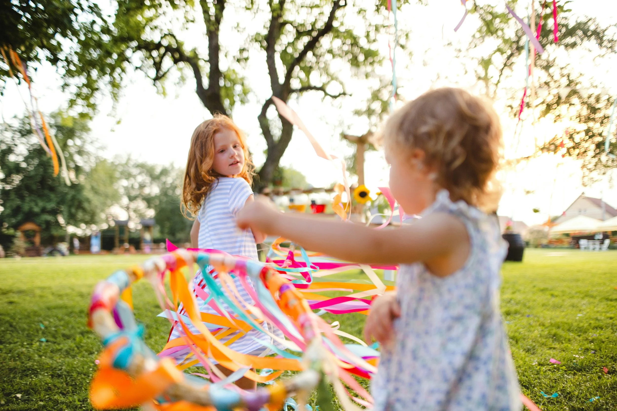 Two young girls playing outdoors with colorful ribbons on a bright sunny day, with trees and a playground in the background.