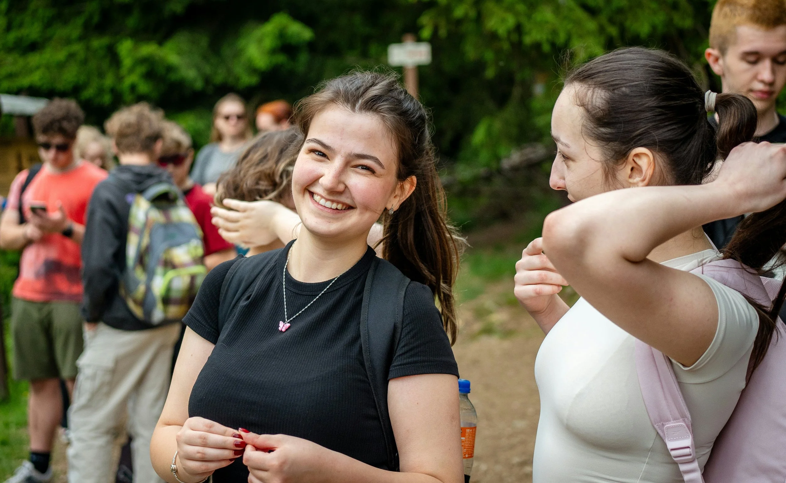 A group of young people outdoors, with two girls in the foreground smiling and talking, others in the background standing and using a cell phone, surrounded by green trees.