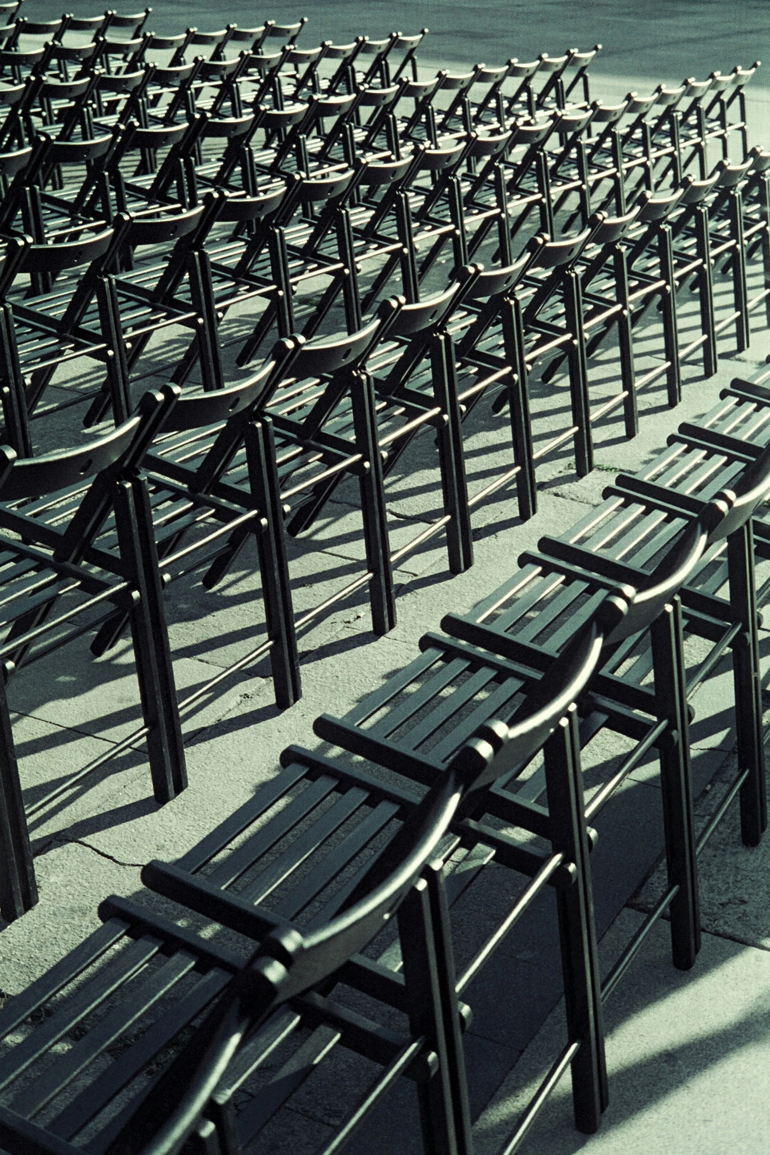 Multiple empty black metal chairs lined up outdoors on a concrete surface, casting shadows from sunlight.