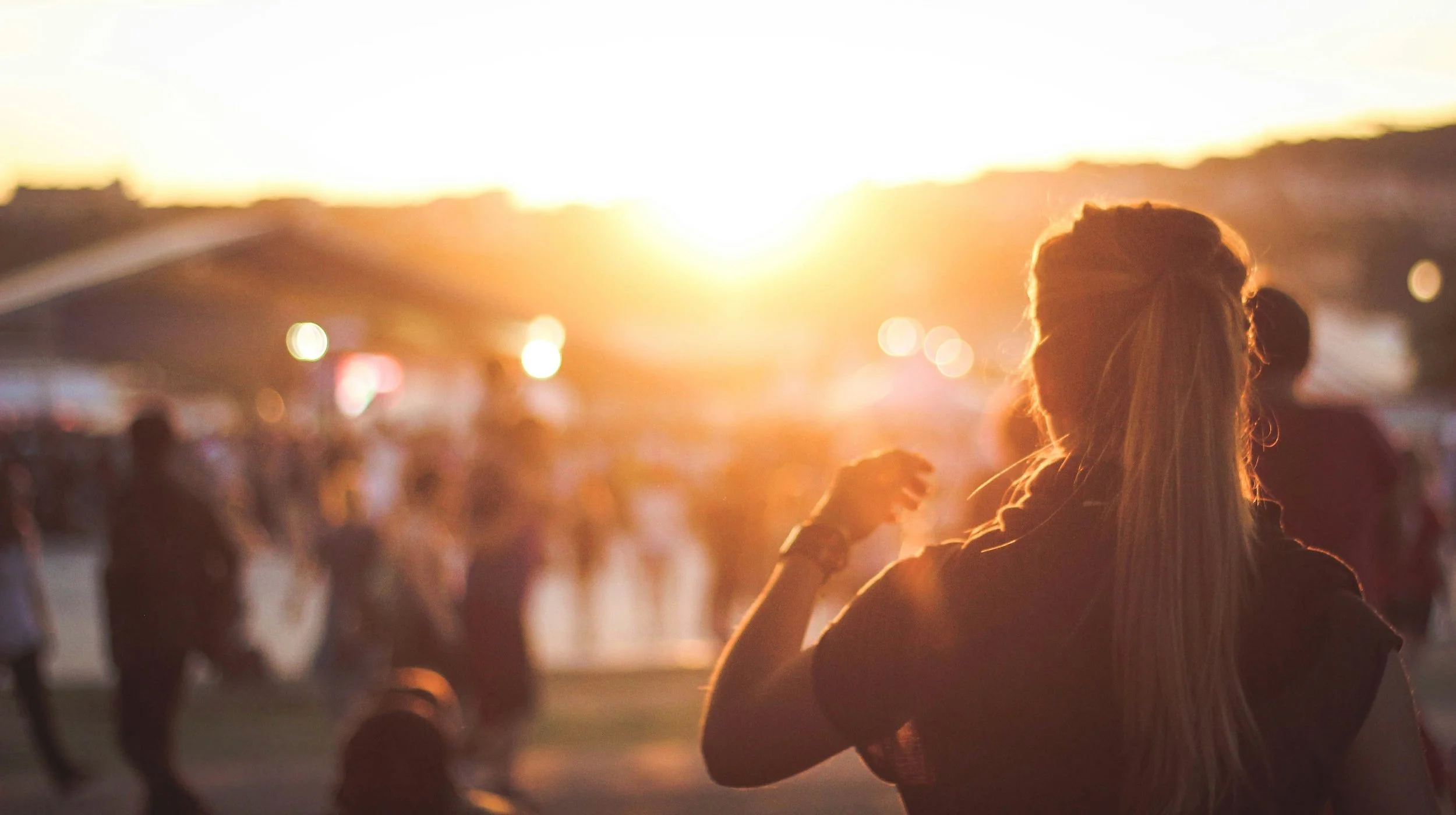 Person with long hair in a bun smiling with a crowd at sunset or sunrise.