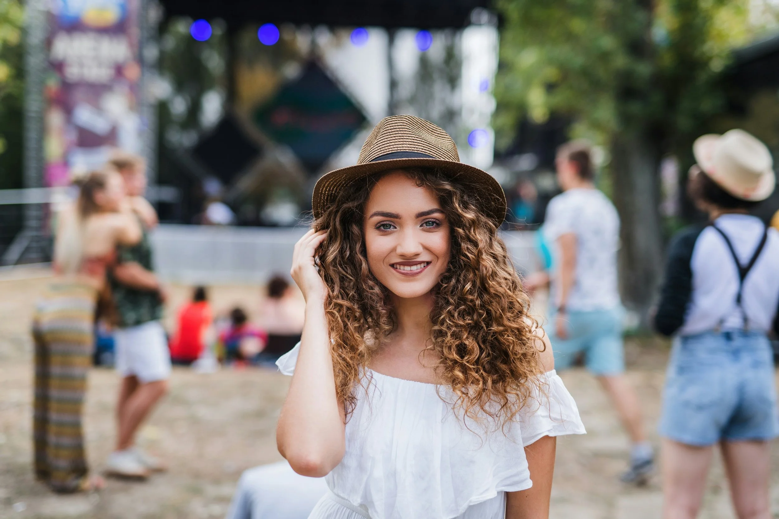 A young woman with curly hair, wearing a white top and a straw hat, smiling and touching her hair at an outdoor event with people in the background.