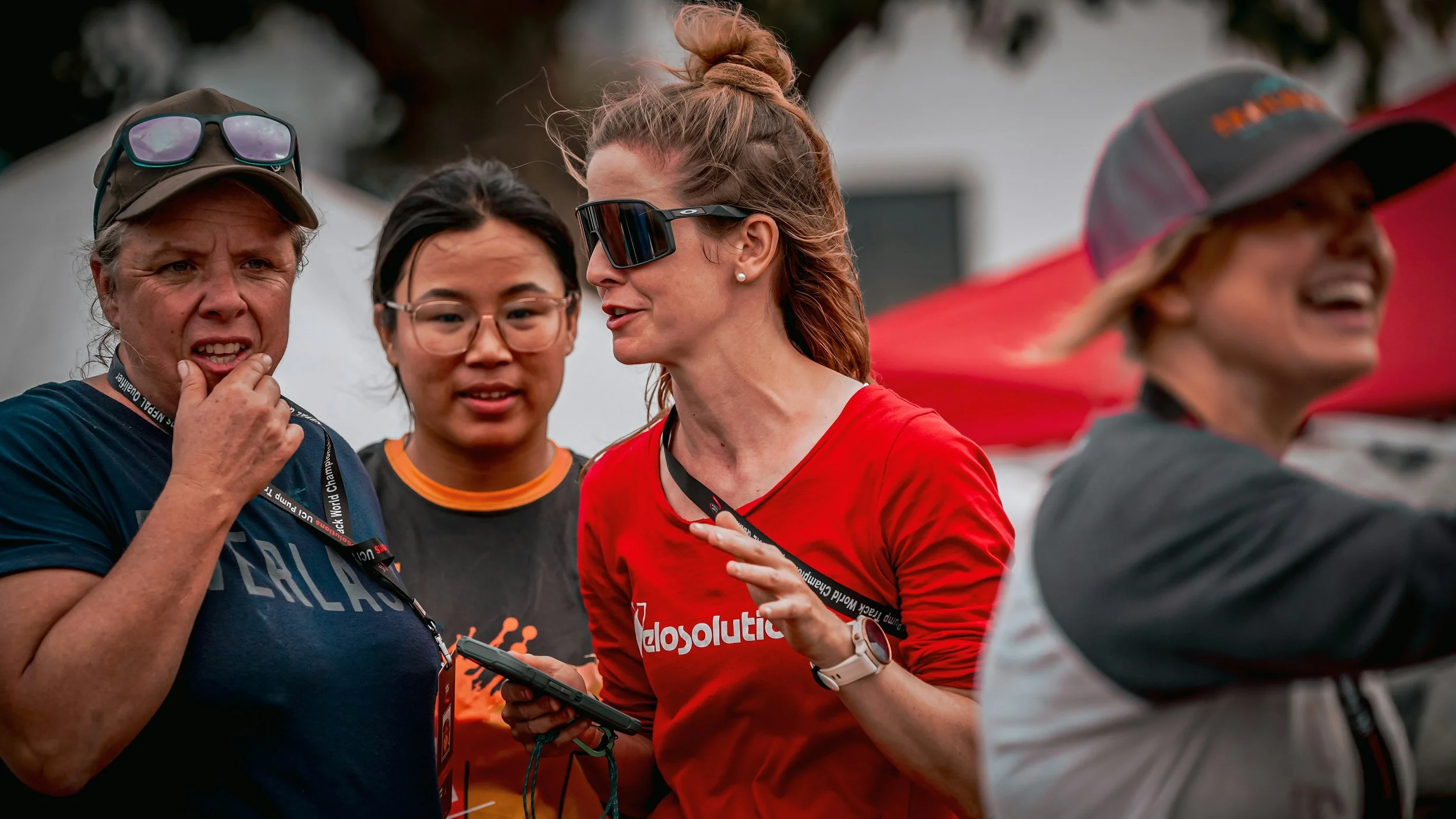 Four women outdoors, talking. One wears sunglasses on her cap, one wears glasses, another has oversized sunglasses and a woman on the far right with a cap and blurred face. They wear athletic shirts and have event lanyards.