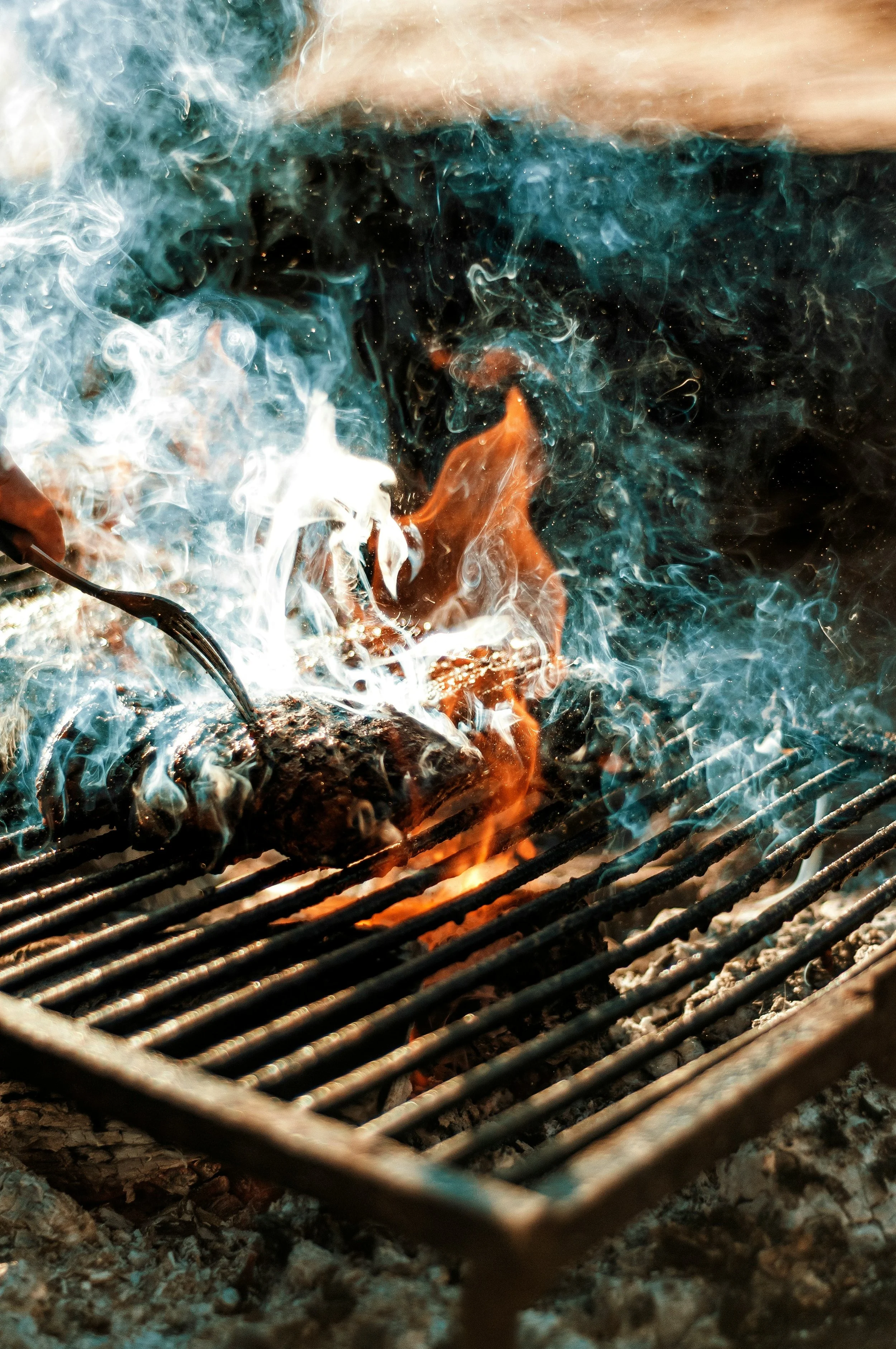 Close-up of a grill with glowing charcoal and burning wood, with white and orange flames and smoke rising.
