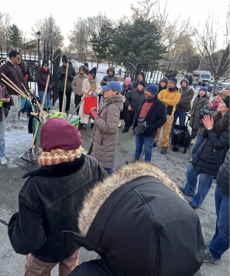 People gathered outdoors in winter clothes listening to a woman speaking at a community event.