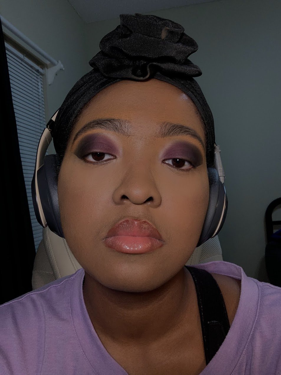 A person with dark skin wearing headphones, a black head wrap, purple eyeshadow, and glossy lips. They are looking directly at the camera.