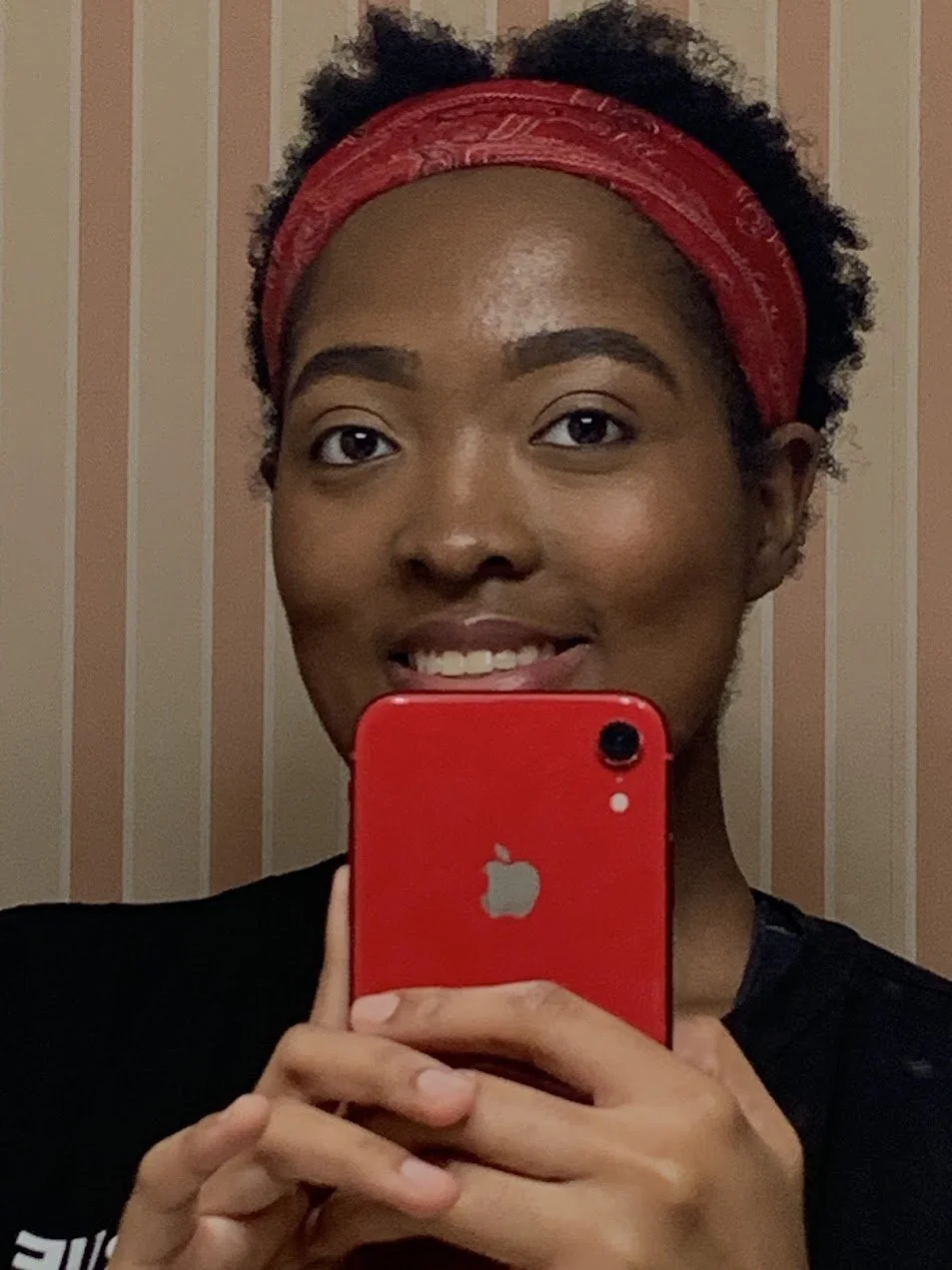 A young woman taking a selfie with a red iPhone, wearing a red headband and smiling, with a striped wallpaper background.