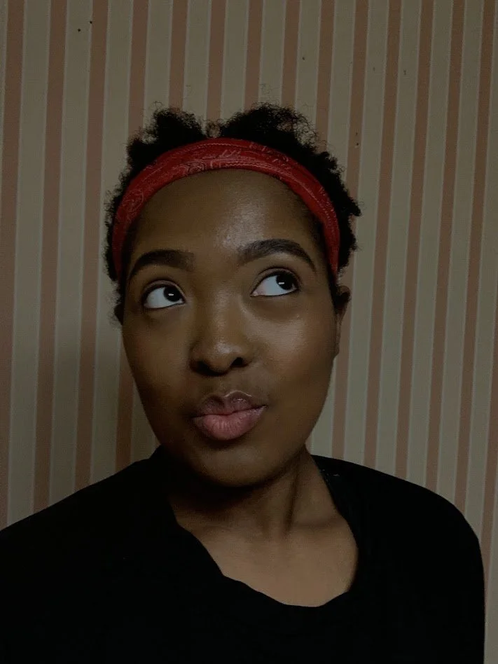 A young woman with dark skin and short curly hair wearing a red headband, looking up and to the side, against a striped wallpaper background.