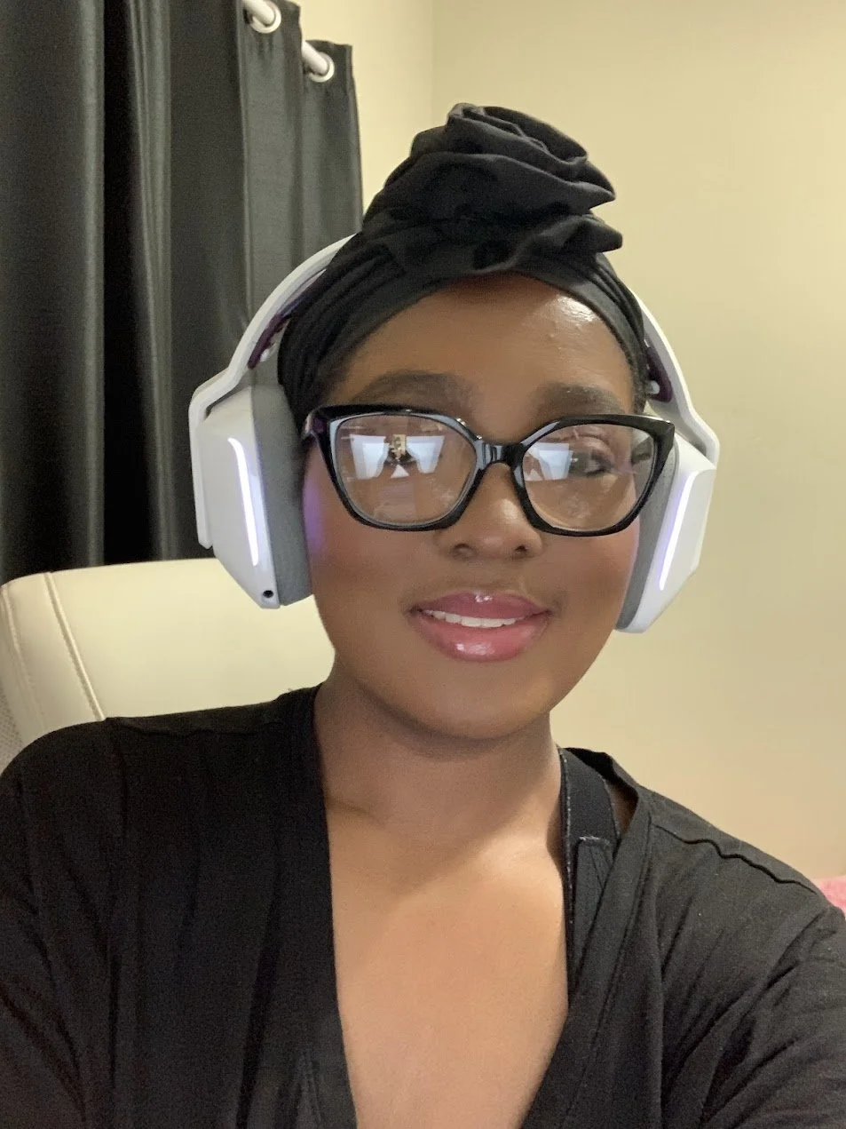 A woman with glasses and a black headwrap wearing white over-ear headphones, smiling in an indoor setting.