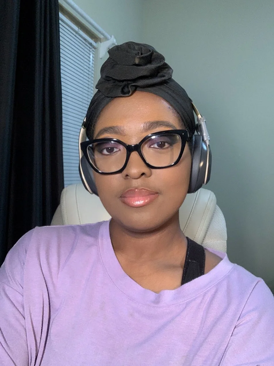 A woman with glasses and a black headwrap is wearing headphones, sitting in a chair, with blinds and a window in the background.