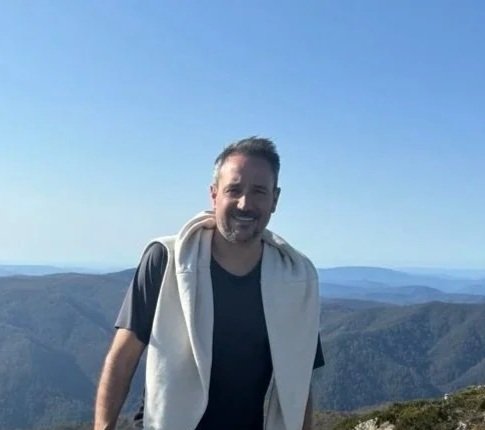 Man standing outdoors on a mountain with a scenic view of distant hills and a clear blue sky. Chris Monahan