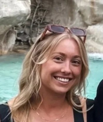 A smiling woman with blonde hair wearing sunglasses on her head, standing outdoors near a body of water with rocks and trees in the background.