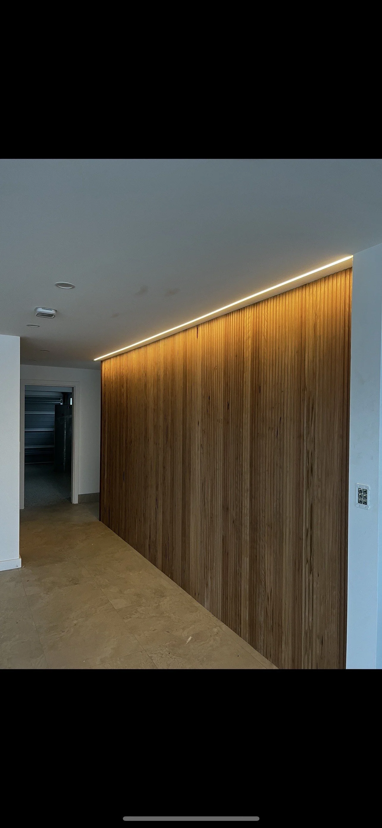 Interior of a modern room with wooden wall paneling, a ceiling LED strip light, and beige tiled flooring, leading to a dark garage or storage area.