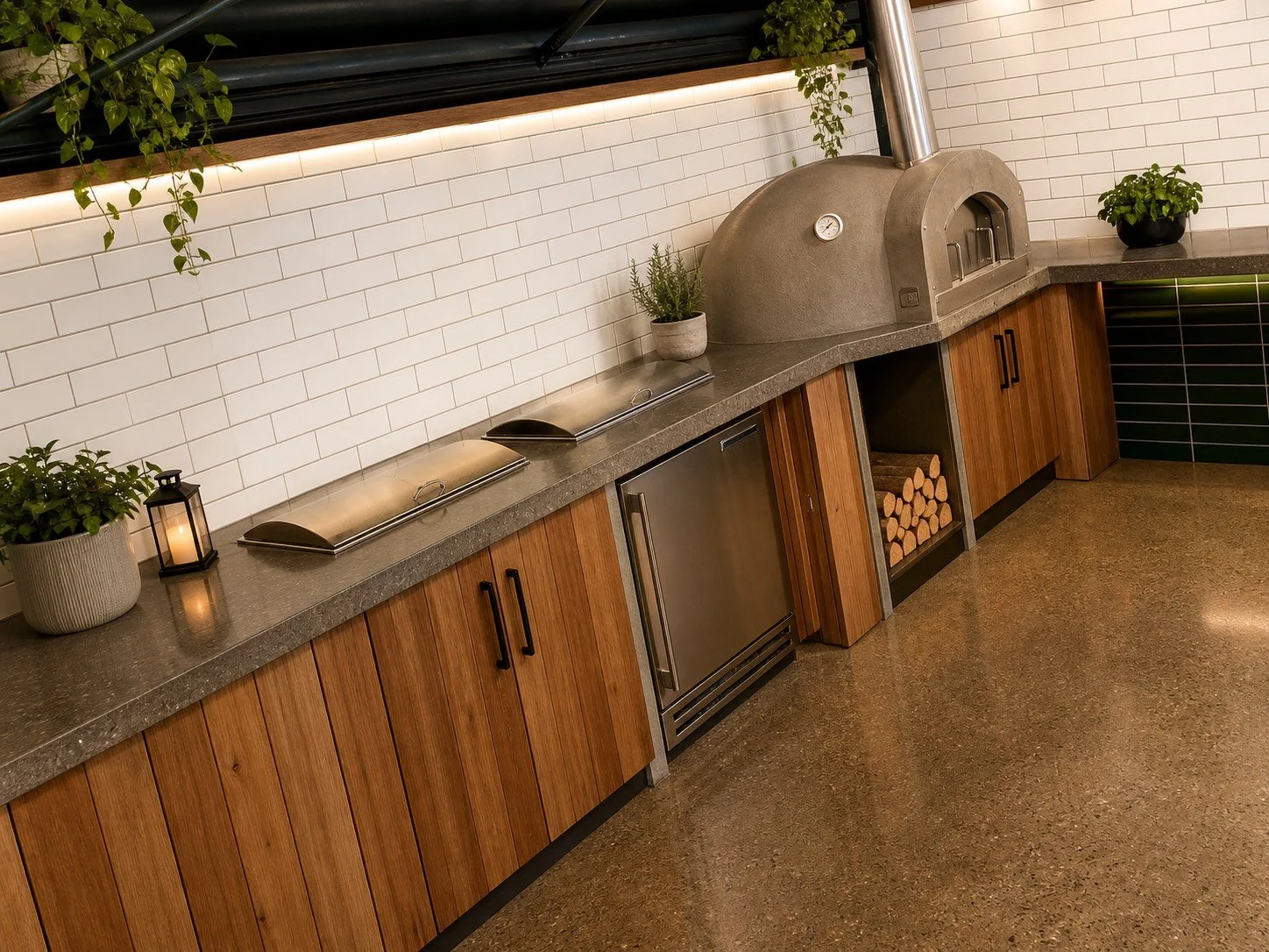 Outdoor kitchen with a wood-paneled countertop, a pizza oven, a small refrigerator, wood storage, plants, and a lit candle.