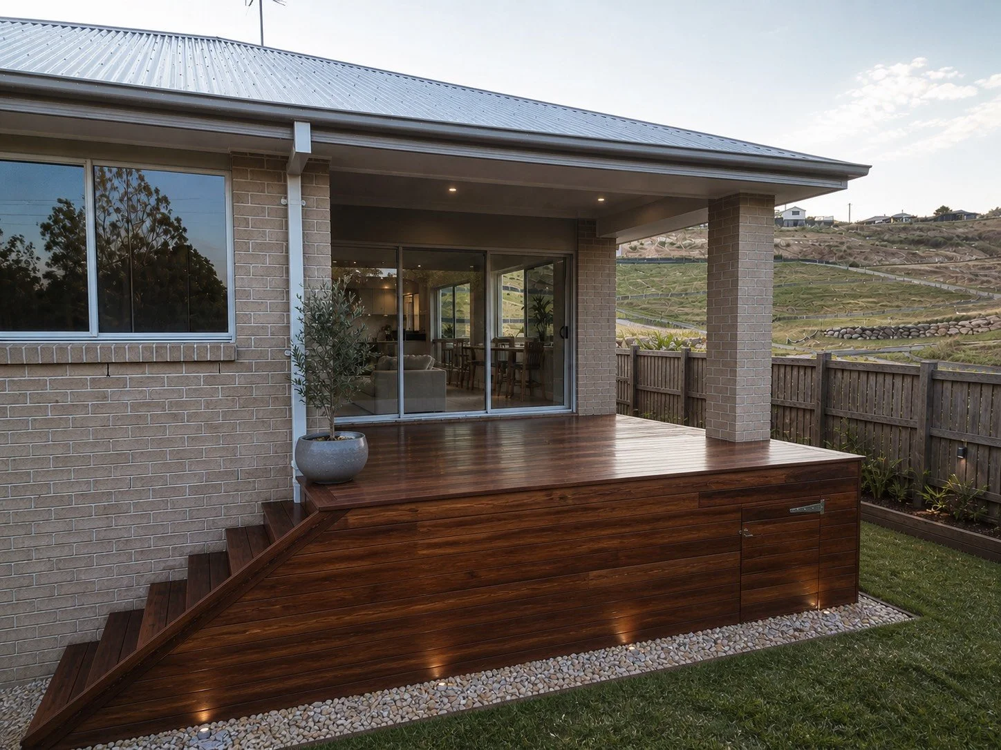 Backyard with a wooden deck and stairs, exterior brick house with large windows and sliding glass door, potted plant, wooden fence, and hilly landscape in the background.