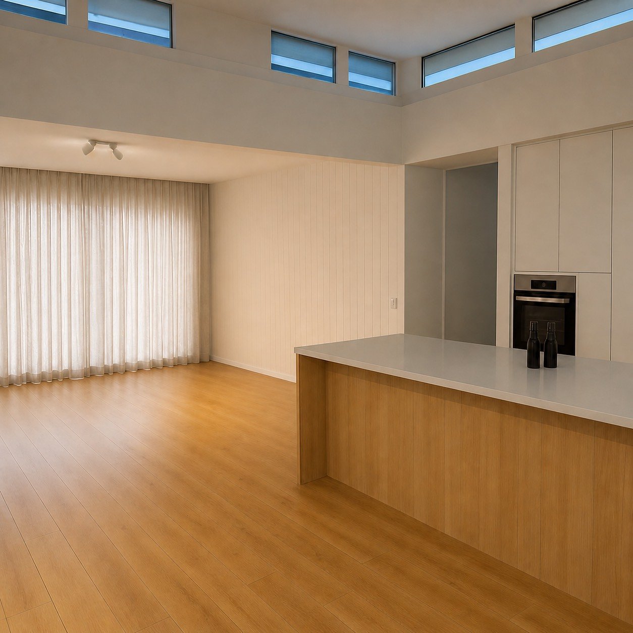 Modern open-concept living space with wood flooring, white walls, a large window with curtains, and a kitchen area with white cabinets, a built-in oven, and a kitchen island with two black bottles on top.