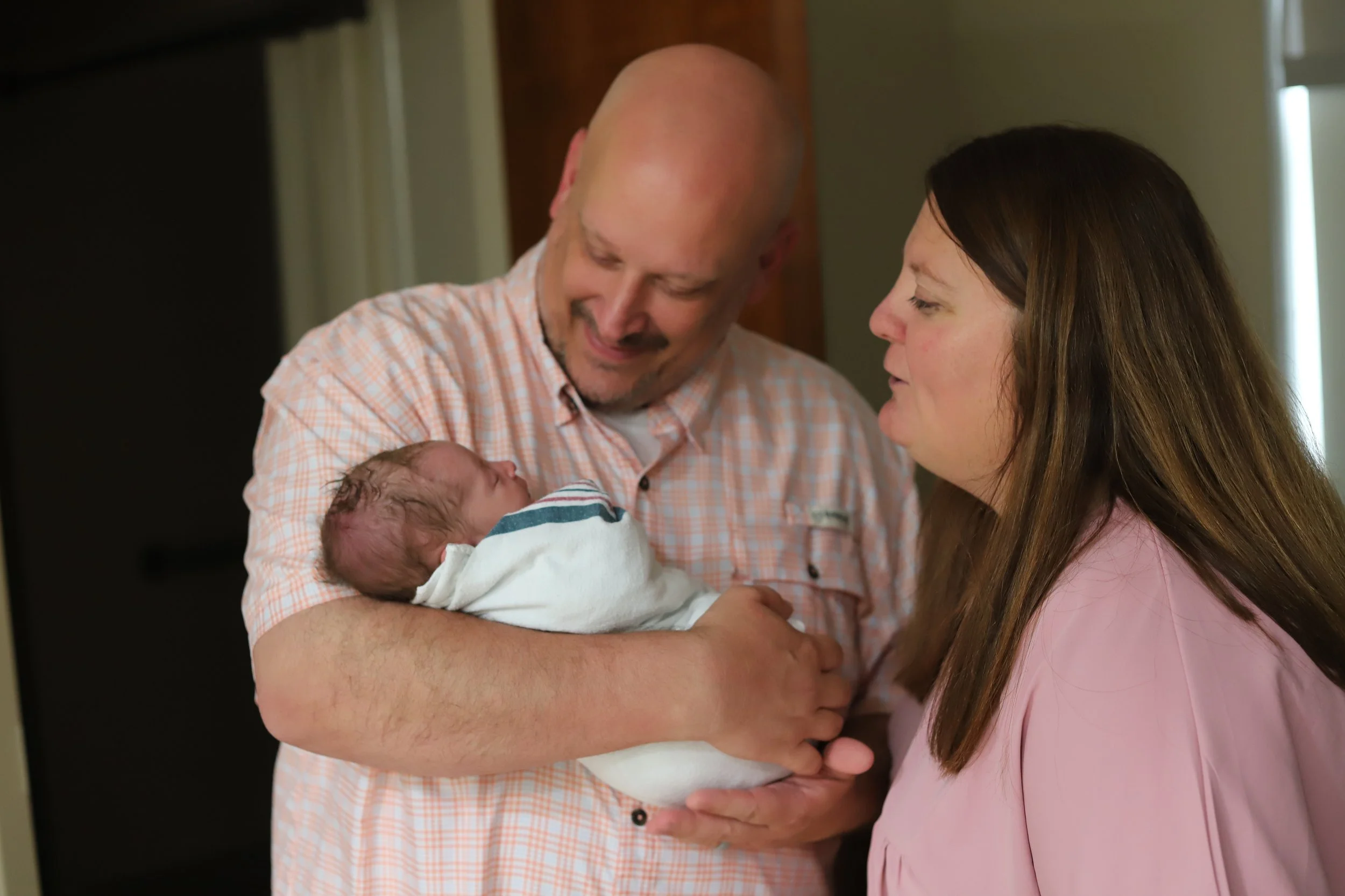 Bald man holding a newborn baby, smiling, with a woman looking at them, indoors.