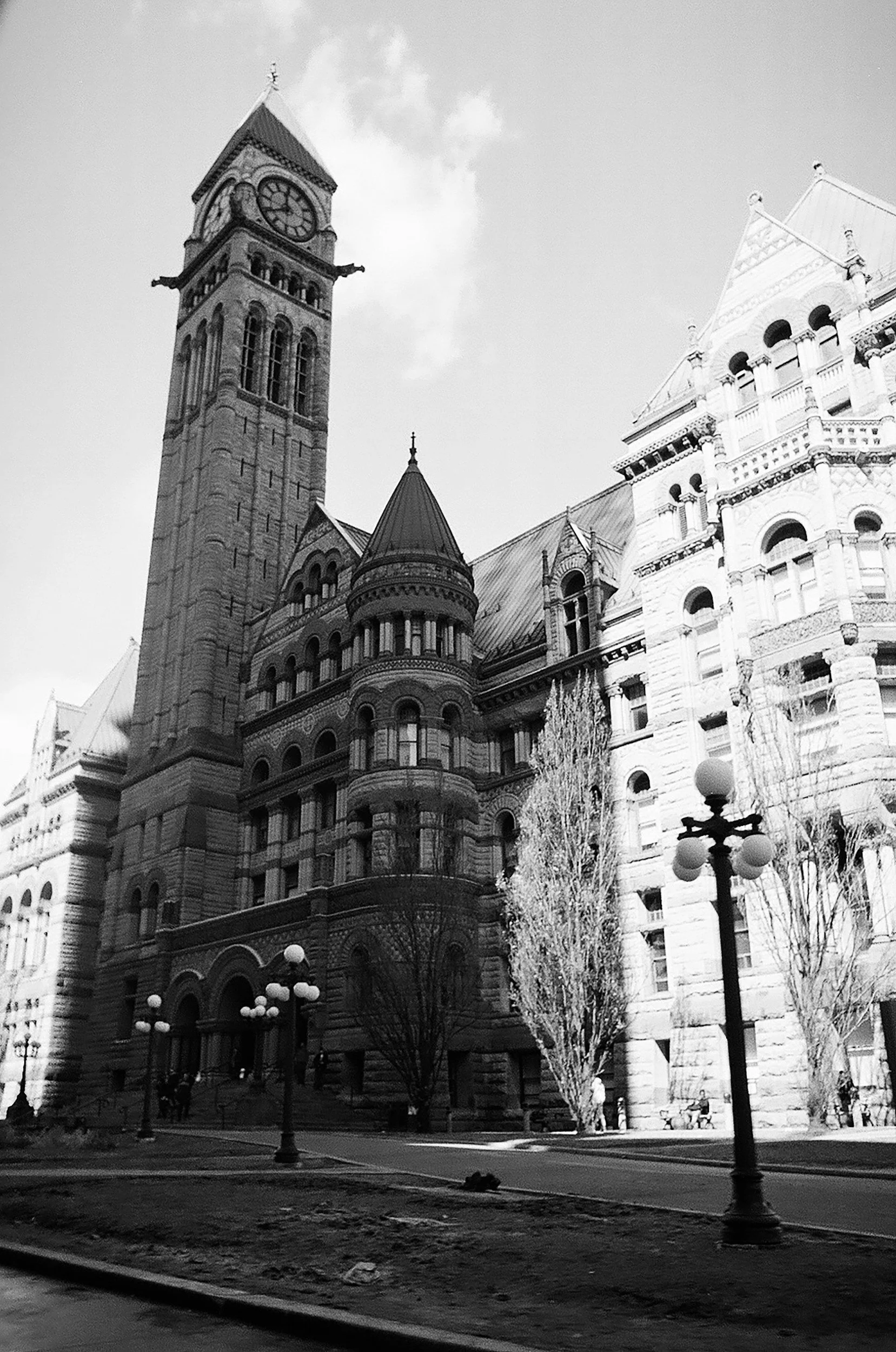Black and white photo of a historic building with a clock tower, ornate architecture, and a pointed roof. Street lamps and leafless trees are in the foreground.