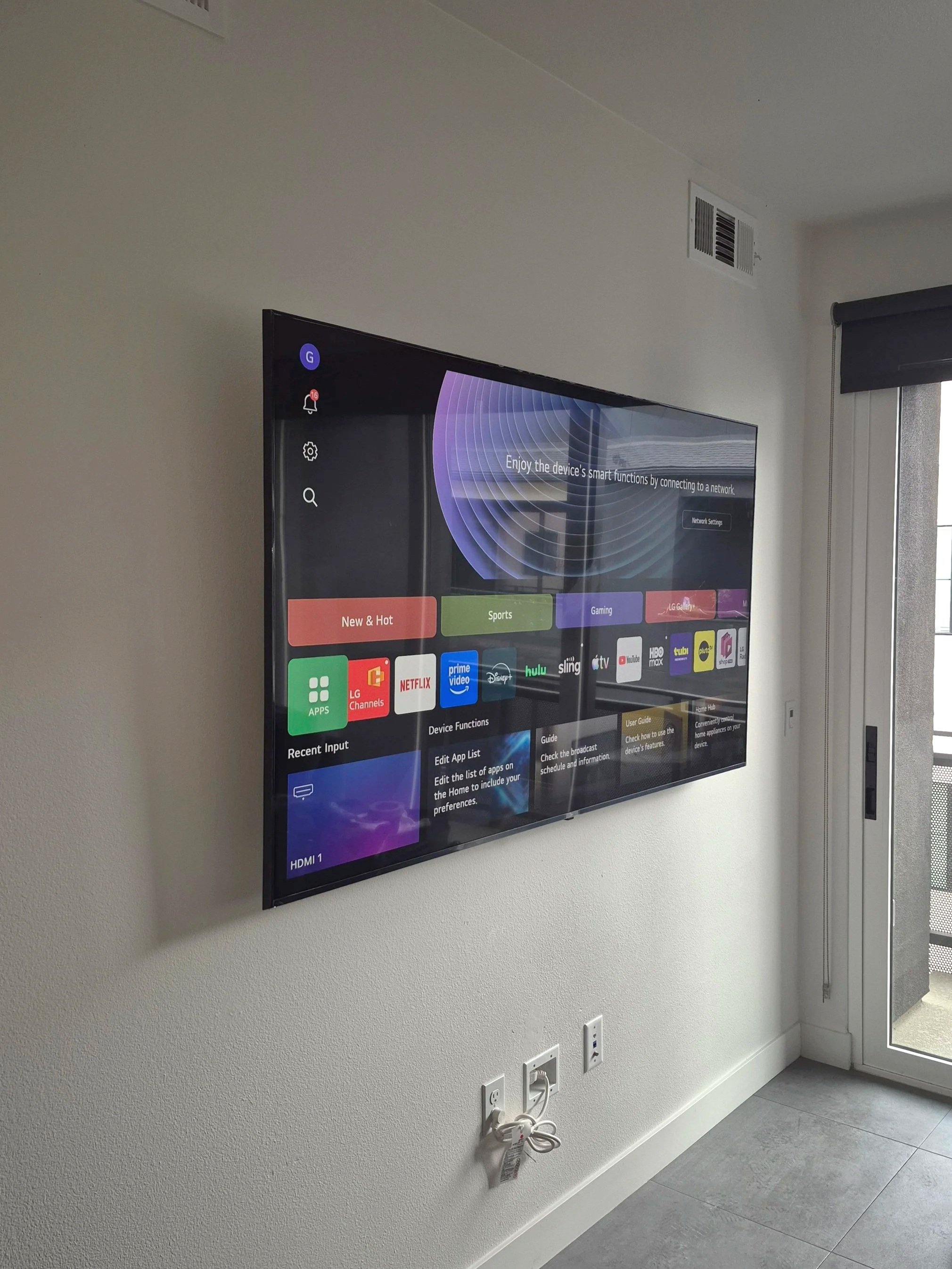 A flat-screen TV mounted on a white wall displaying a smart TV menu with app icons such as Netflix, Hulu, Disney, and YouTube, located near a window with a black roller shade and visible electrical outlets on the wall below.