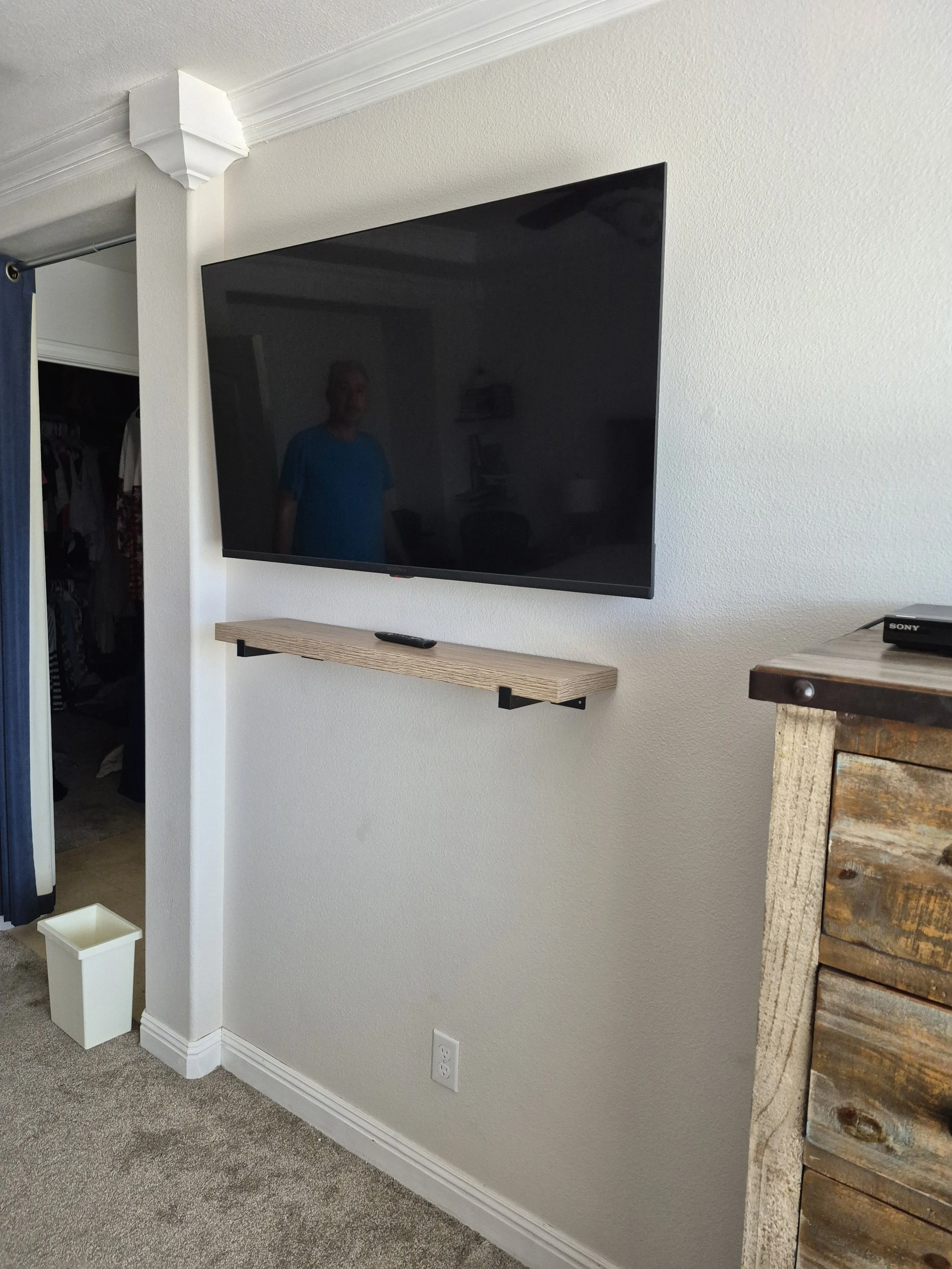A flat screen TV mounted on a white wall with a small wooden shelf beneath it. There is a person reflected on the TV screen, and a remote control on the shelf. To the left, there is a doorway with a closet visible in the background. A small white trash bin is on the floor near the wall, and part of a rustic wooden piece of furniture is visible on the right.