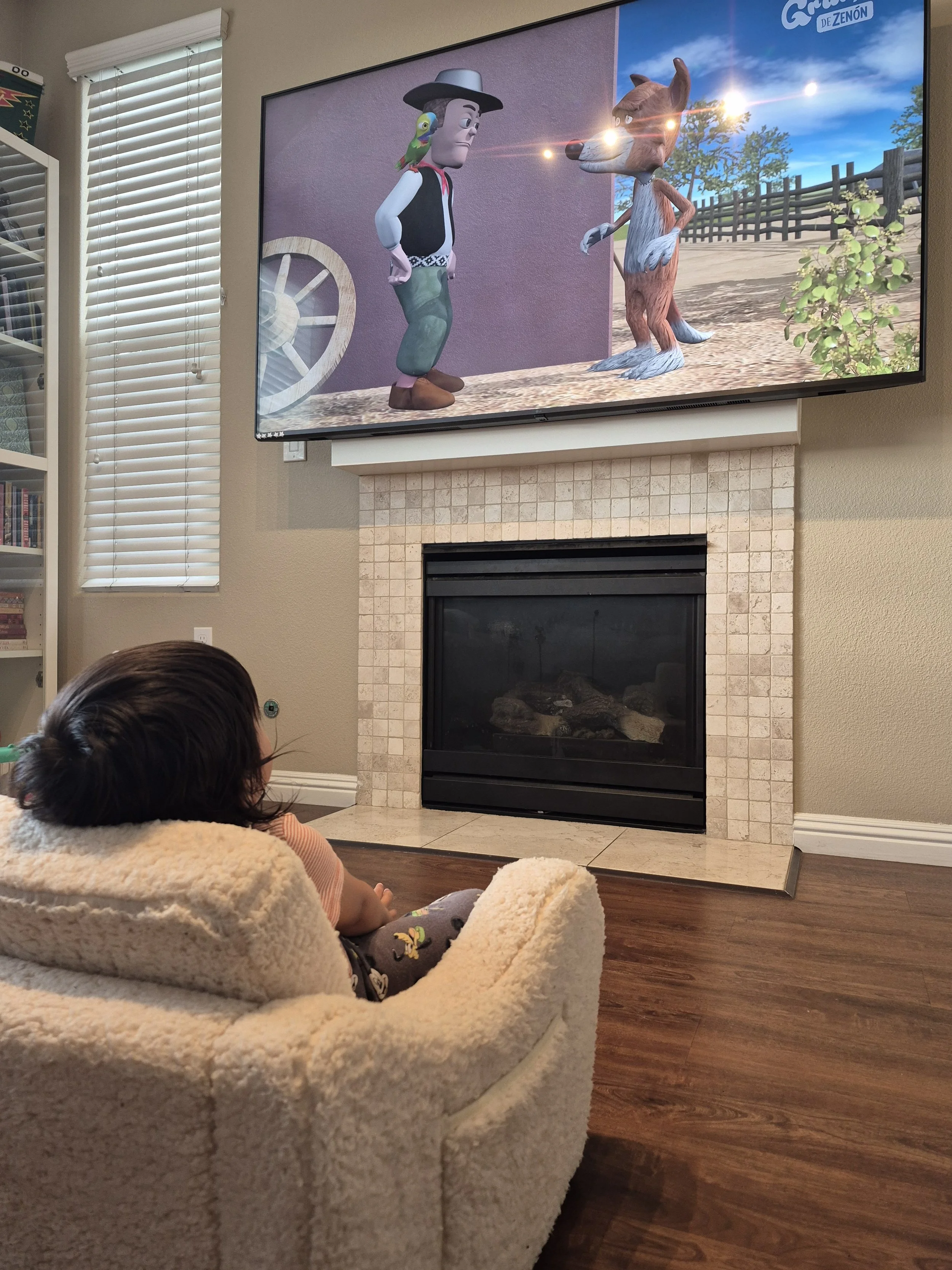 A young child with dark hair playing on a plush chair in a living room while watching an animated show on a large wall-mounted TV. The TV displays a cartoon scene of two characters: a cowboy with a parrot on his shoulder and a wolf with a fence and a bright sky in the background.
