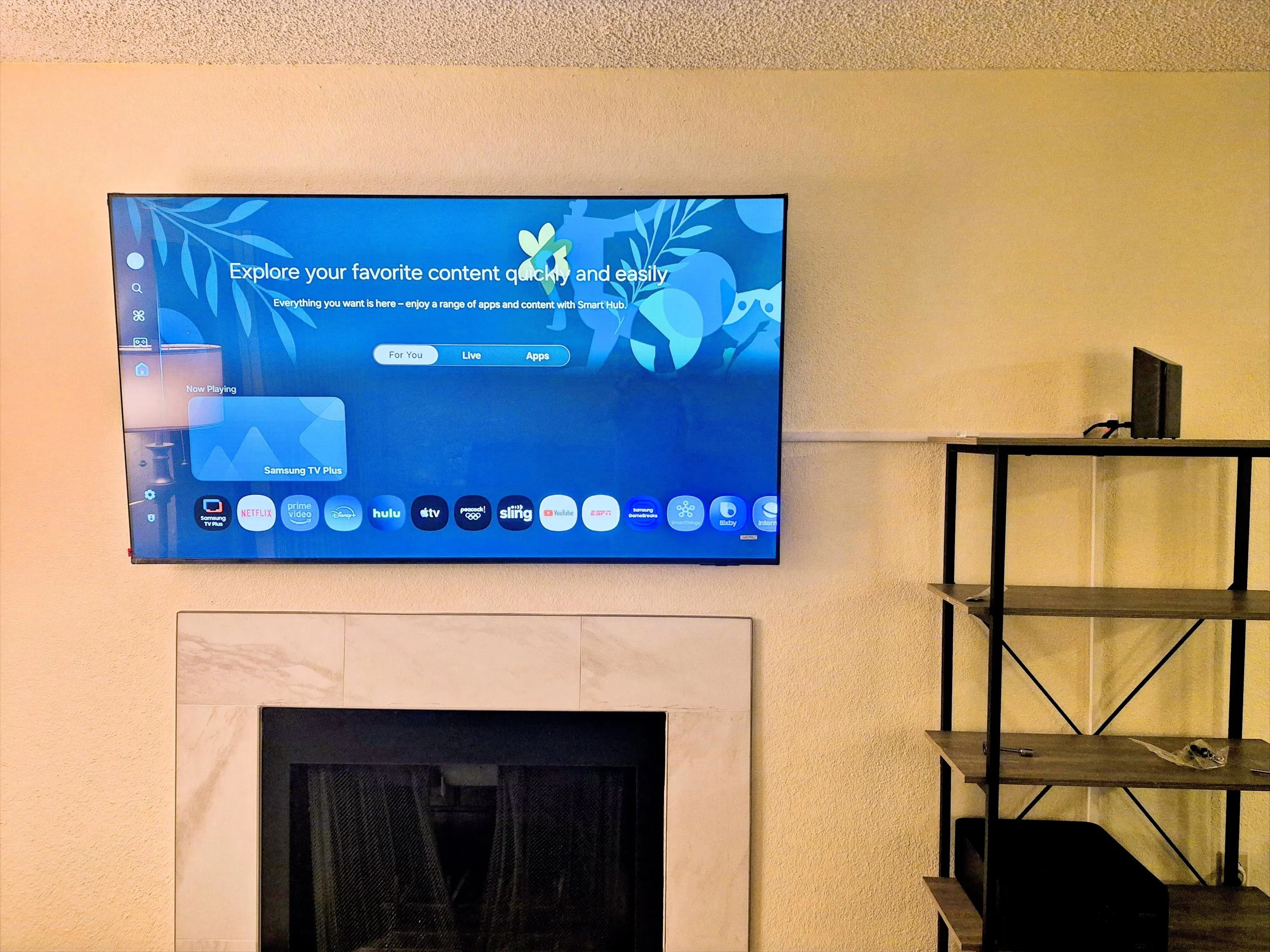 Photo of a flat screen TV mounted on a beige wall, showing a streaming service interface. To the right, there is a black, four-tier metal shelving unit. Below the TV, there's a fireplace with white marble surround and a black interior.
