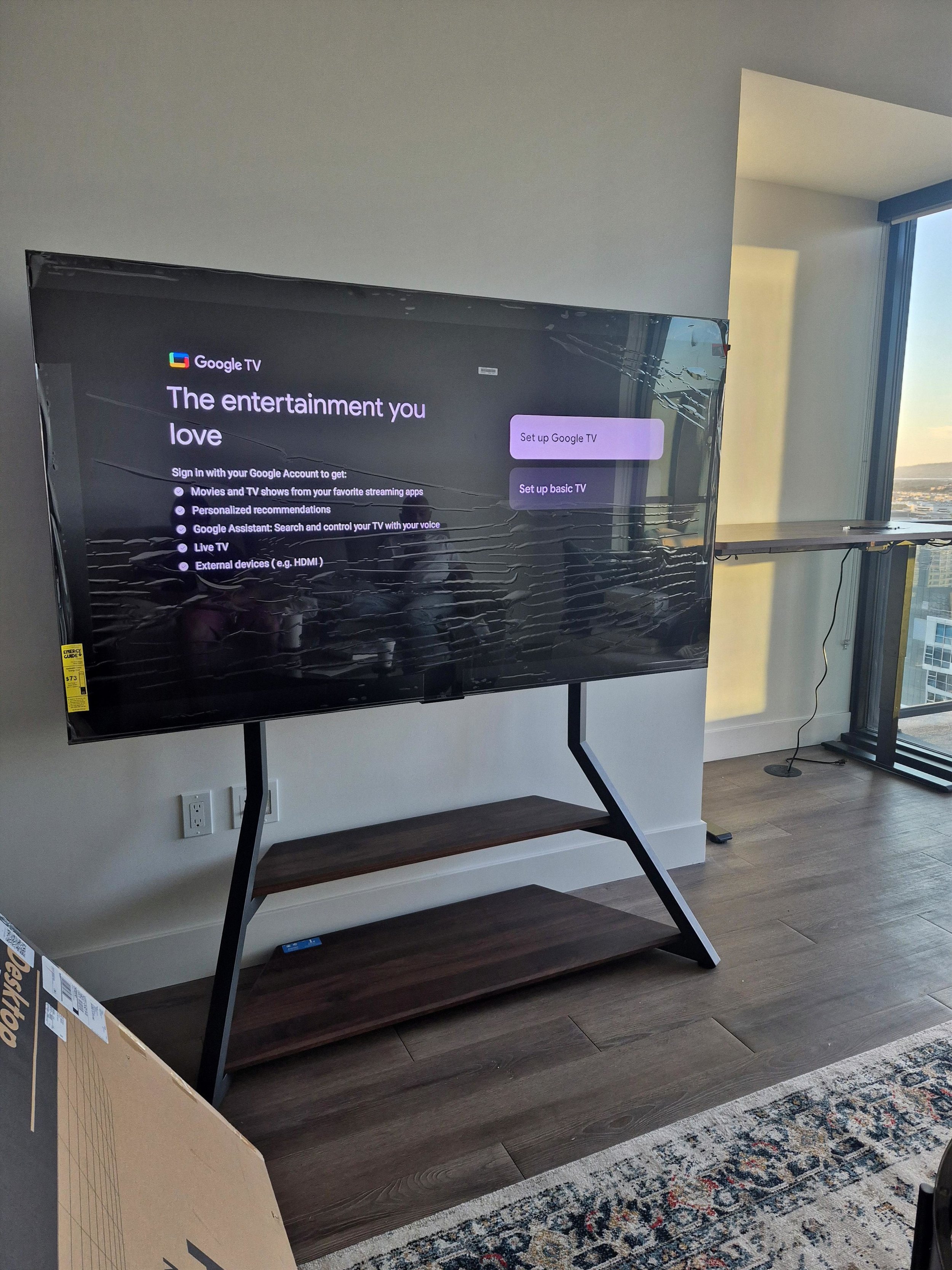 A flat-screen television displaying setup options, placed on a black stand with a wooden shelf, in a room with hardwood flooring and a light-colored rug near a large window with a view of city buildings.