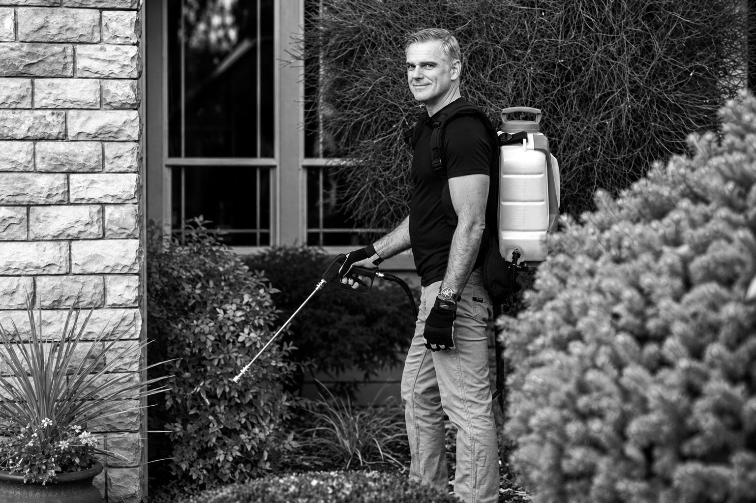 A man with light hair wearing a black t-shirt, light-colored pants, and gloves, holding a weed trimmer outdoors near a house with a brick wall and windows, surrounded by bushes and plants.