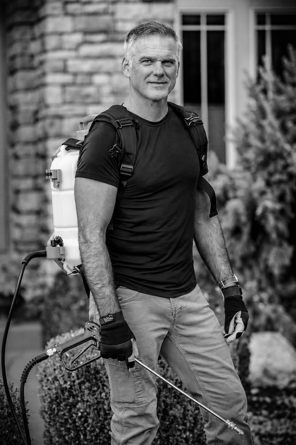A man with gray hair and a beard standing outdoors, wearing a black t-shirt, light-colored pants, gloves, and a wristwatch, carrying a backpack with a spray tank, holding a weed sprayer wand.