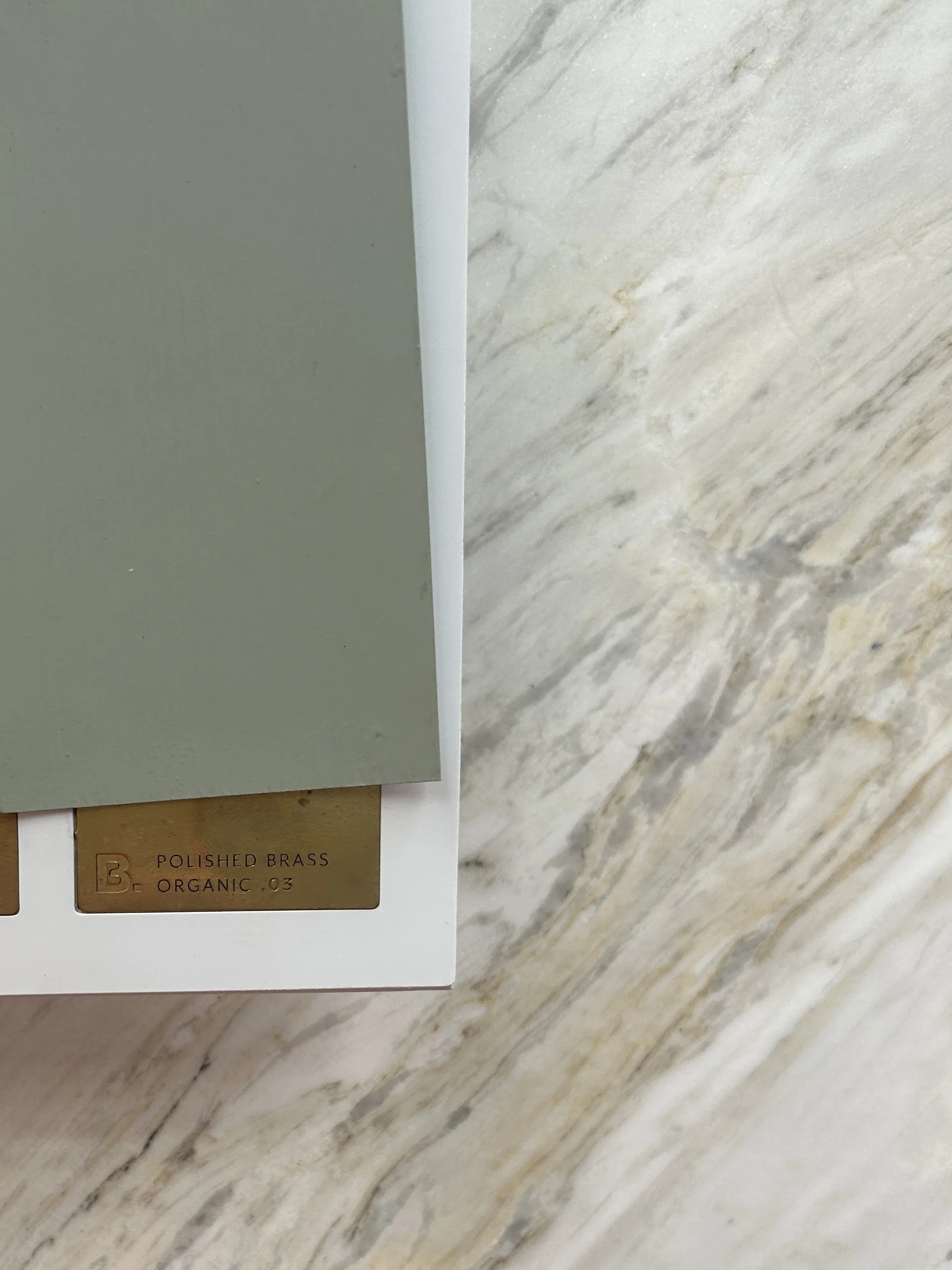 Close-up of a color sample card showing a dark green color and a marble-patterned surface, with a brass sample label reading 'Polished Brass Organic .03'.