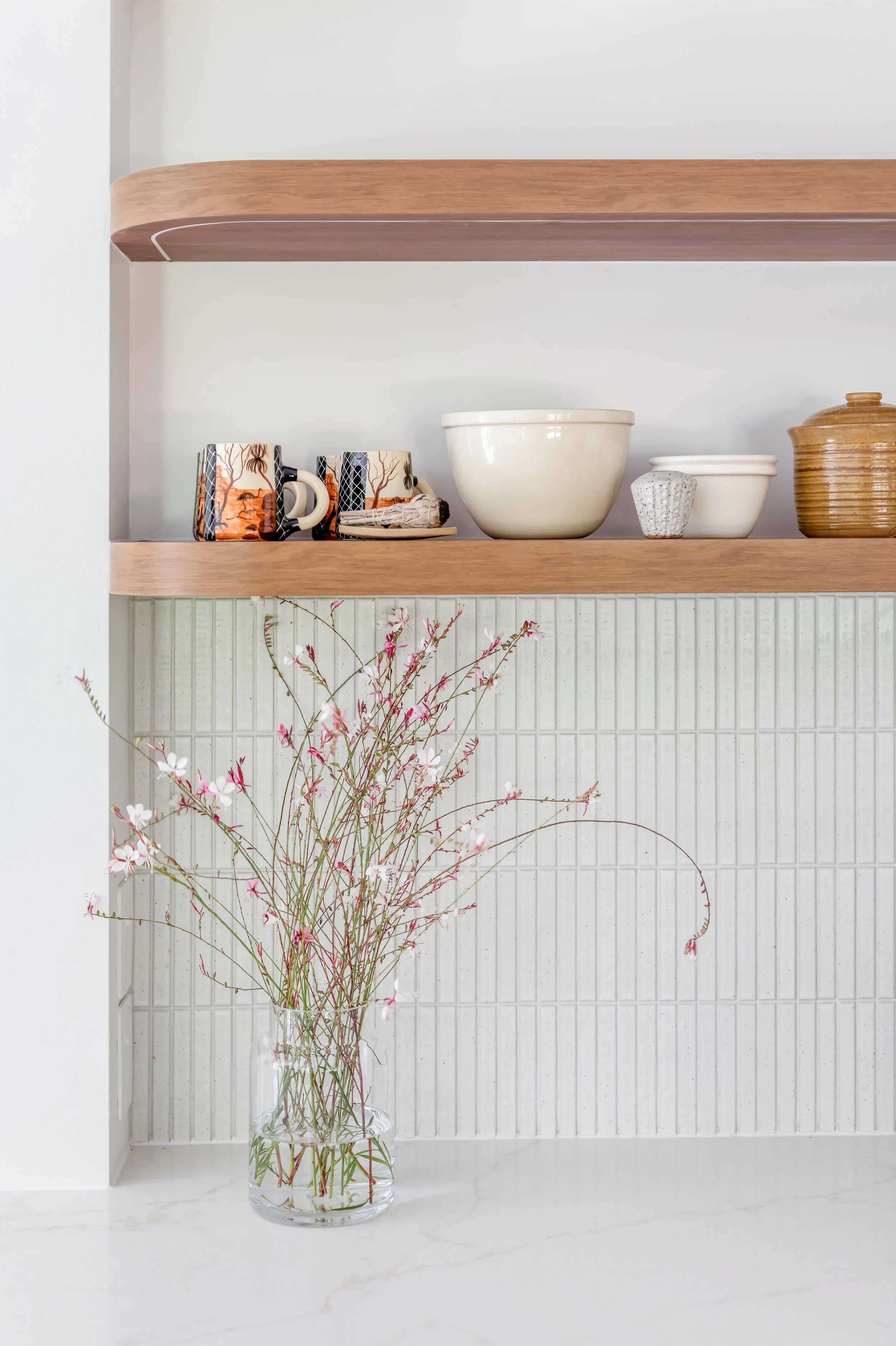 A clear glass vase filled with pink and white flowers with long, thin stems in front of a white wall and a wall-mounted wooden shelf holding various ceramic and wooden containers.