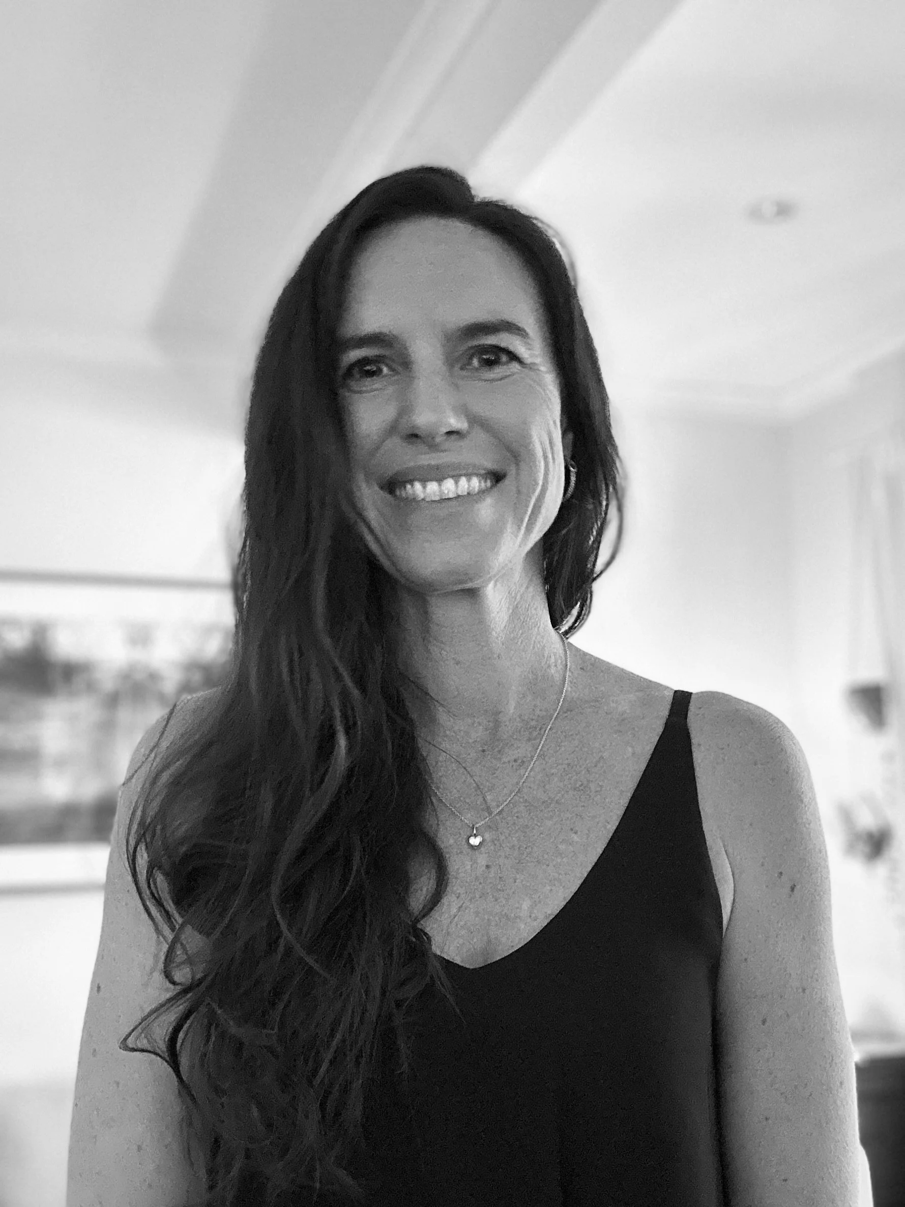 A smiling woman with long dark hair wearing a black top with one strap, a necklace with a heart-shaped pendant, and earrings, standing indoors.