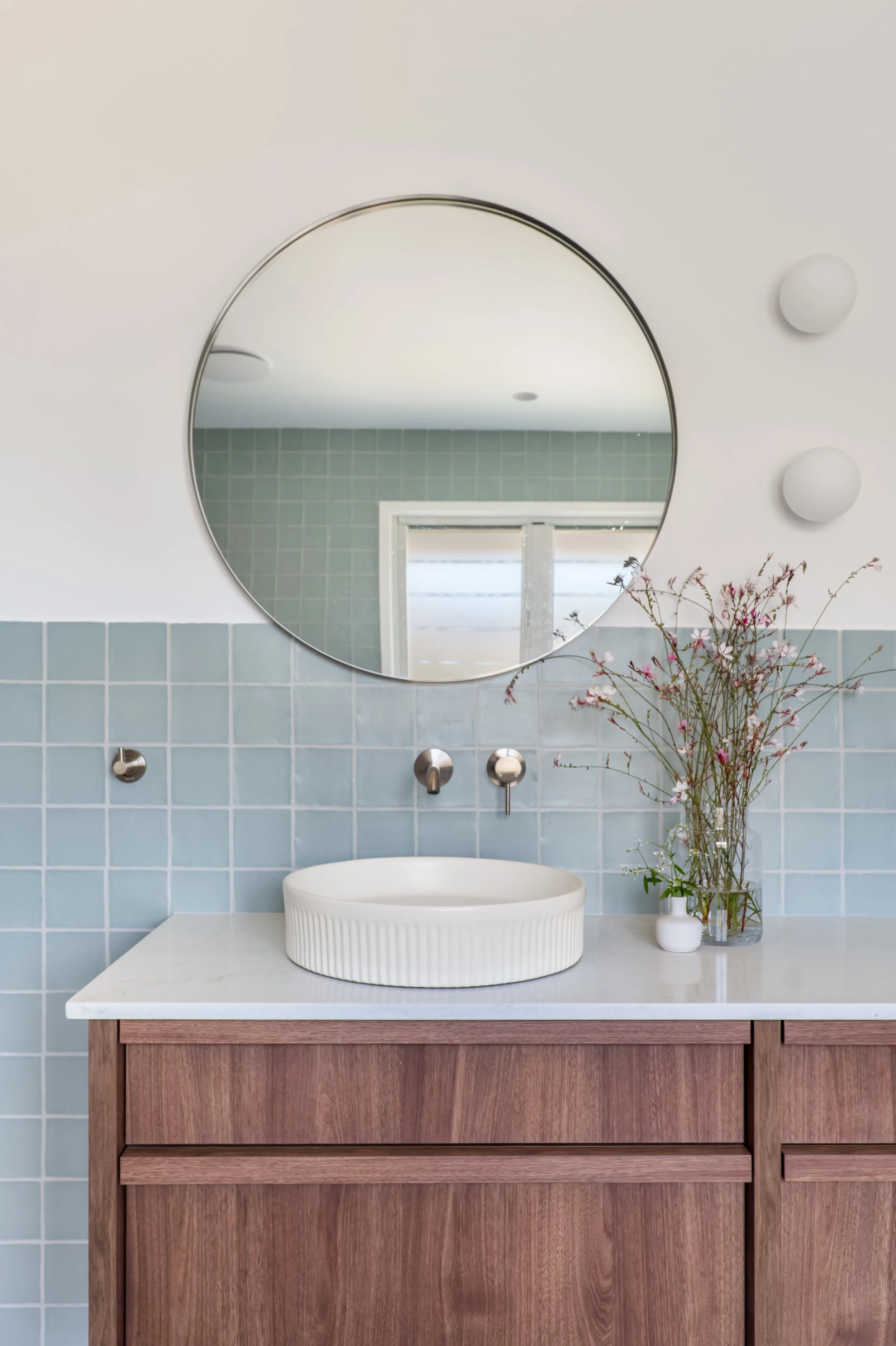 Bathroom vanity with a round vessel sink, a large mirror, blue tiled wall, and a countertop with a vase containing pink and white flowers.