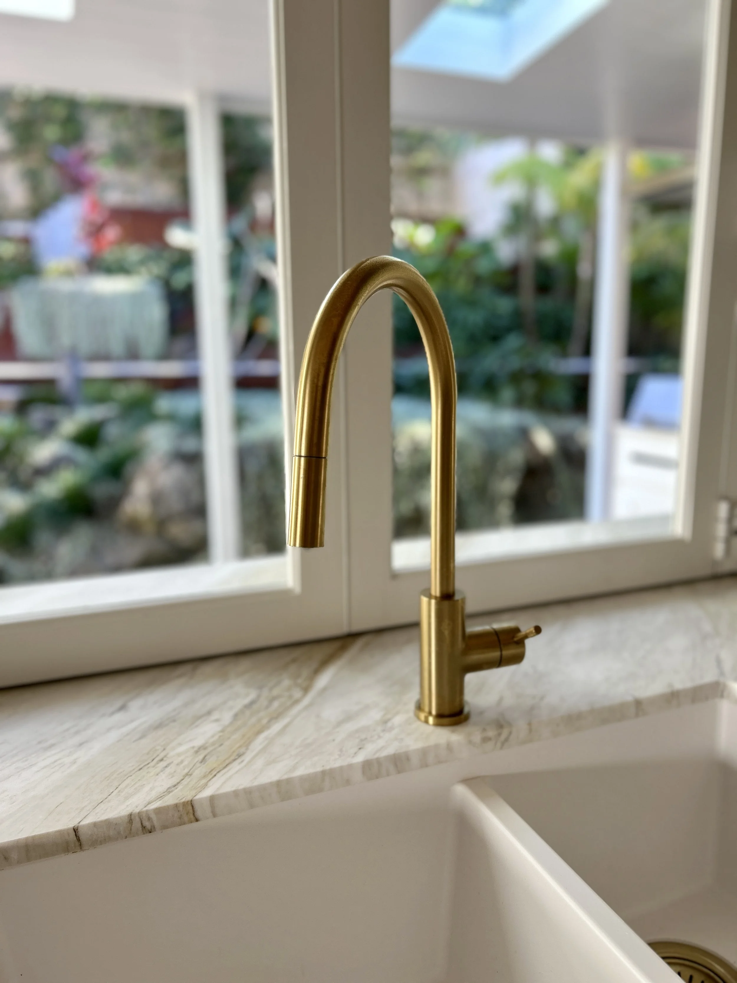 A close-up view of a gold kitchen faucet mounted on a marble countertop in front of a glass window, overlooking a garden.