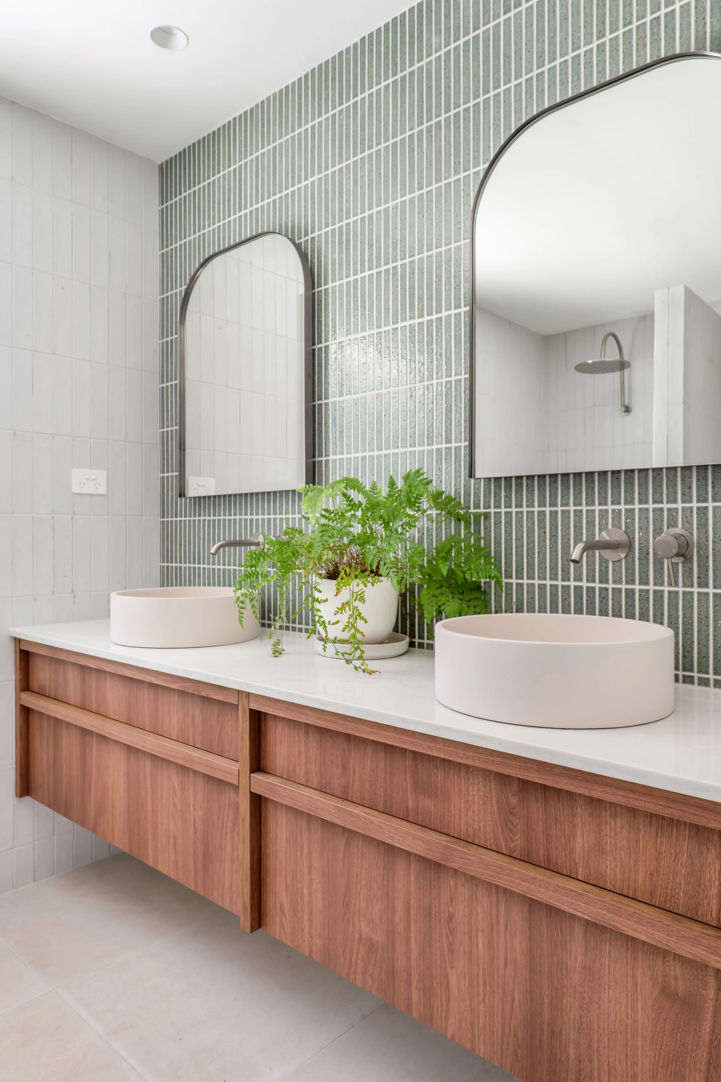 Modern bathroom vanity with two round vessel sinks on a white countertop, wooden cabinet, large mirrors, green plant, and shower with rain shower head in the background.