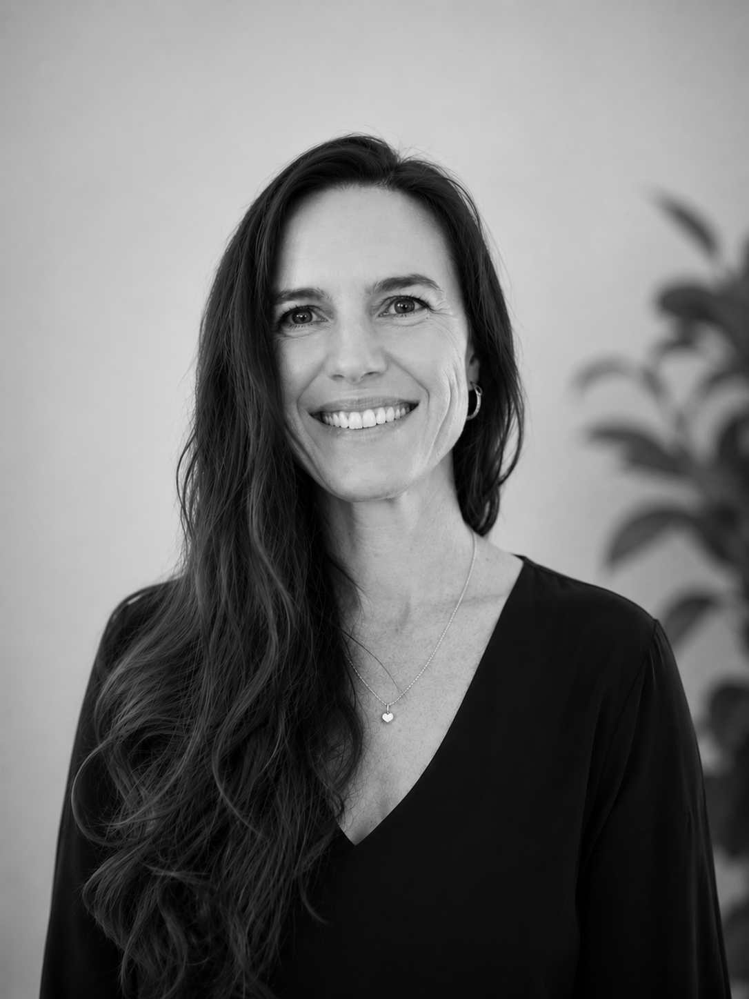A black-and-white portrait of a woman with long wavy hair, smiling and wearing a necklace and a black top, standing in front of a plain background with a blurred plant on the right side.