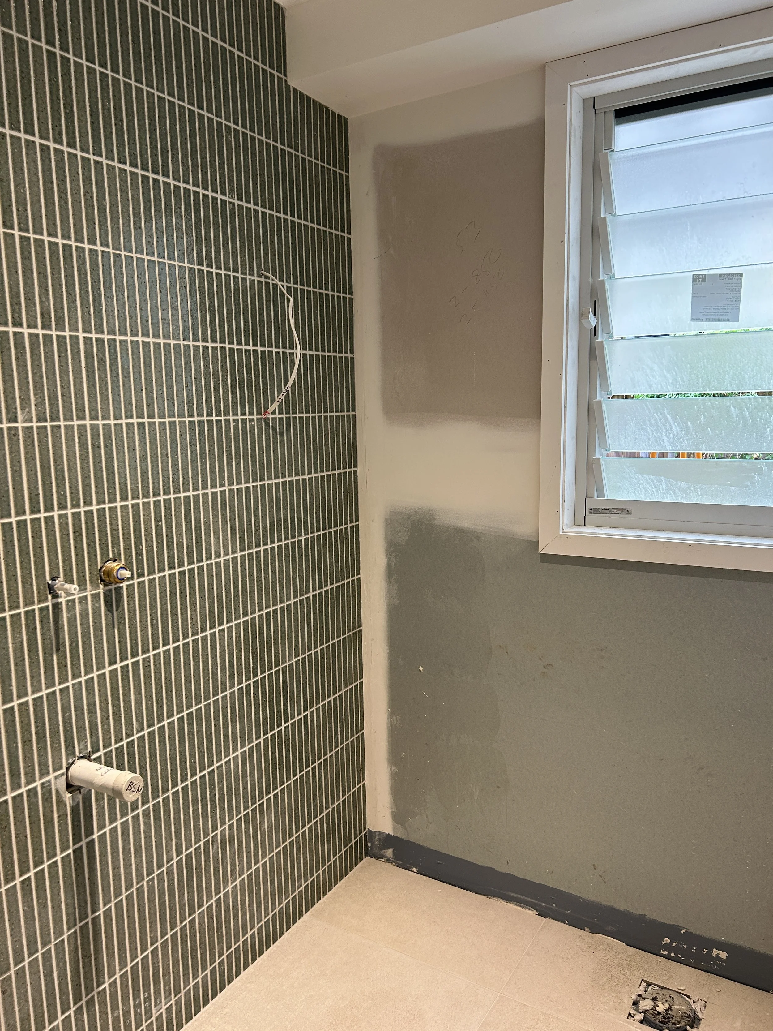 A room under construction with green tile wall on the left, an unpainted wall with markings in the middle, and a window with partially open blinds on the right. The floor has light-colored tiles, and there is a hole in the floor near the window.