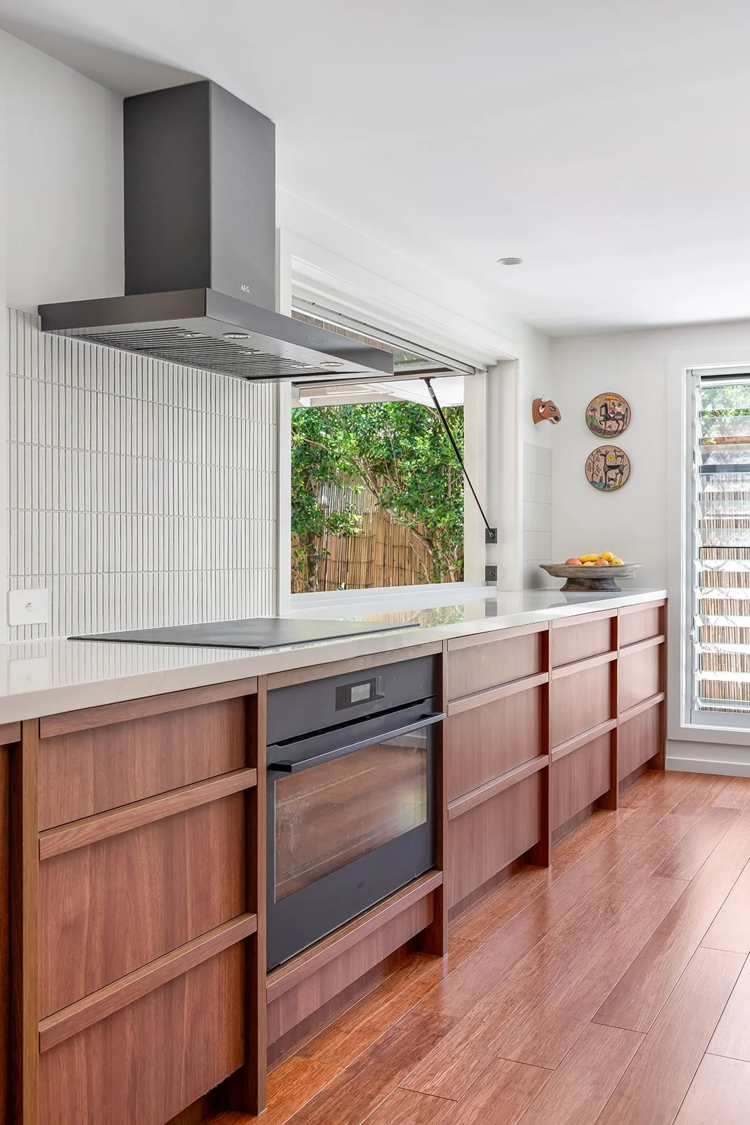 Modern kitchen with wooden cabinets, white countertop, black oven, and range hood. A window shows a garden view, and decorations include three colorful plates on the wall and a bowl of fruit.