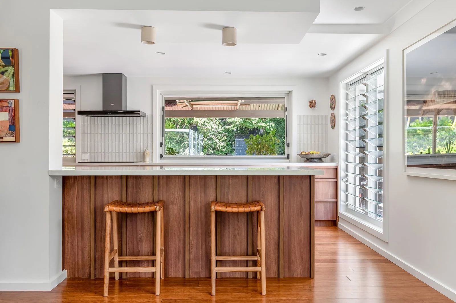 Modern kitchen with wooden bar stools, open window overlooking green outdoor area, white walls, and minimal decor.