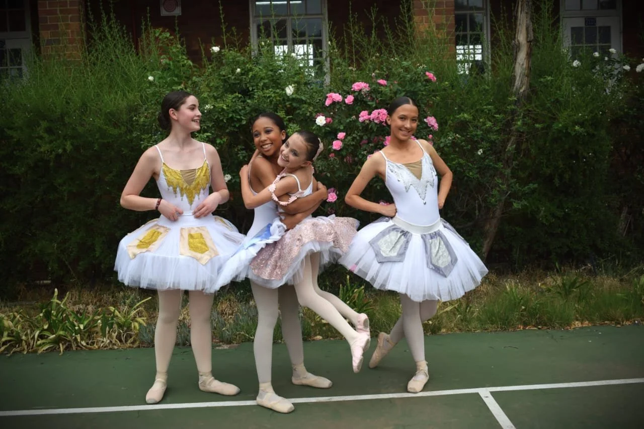 Four young ballet dancers in tutu costumes standing outdoors near greenery and pink roses, smiling and posing for a photo.