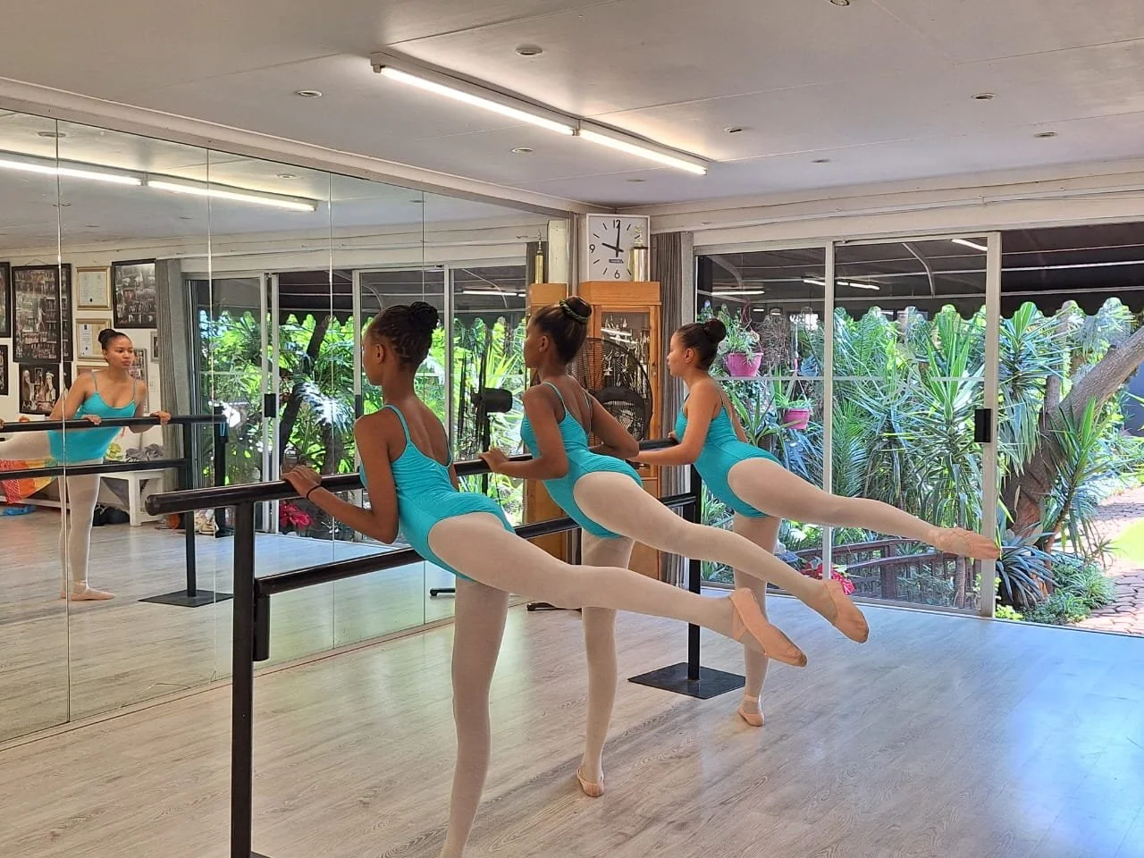 Three young ballet dancers practicing at the barre in a dance studio with large mirrors and a view of green plants outside, with a ballet instructor observing.