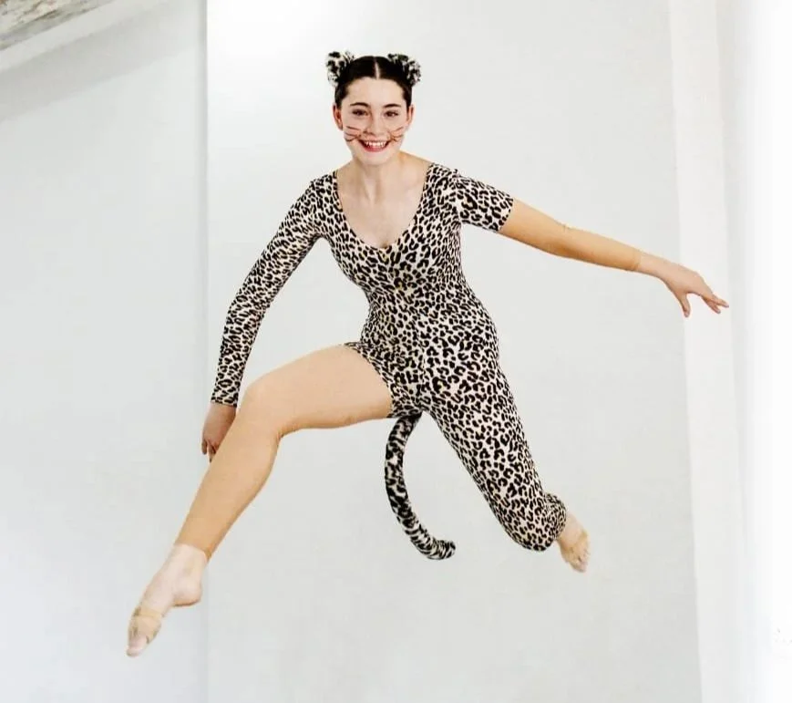 A woman in a leopard print leotard has a cat tail and is jumping in the air with a white wall background.